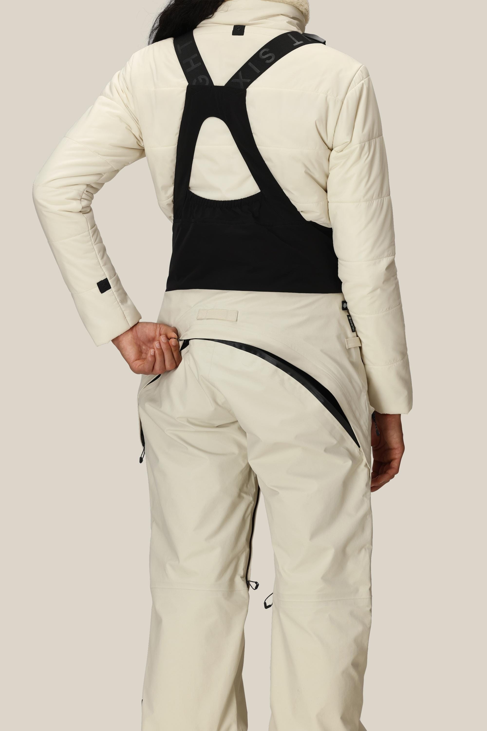 A person wearing the 686 Women's GORE-TEX Dispatch™ Shell Bib in cream, along with a matching jacket featuring a black back panel, is facing away and unzipping the large horizontal rear zipper of the bib.