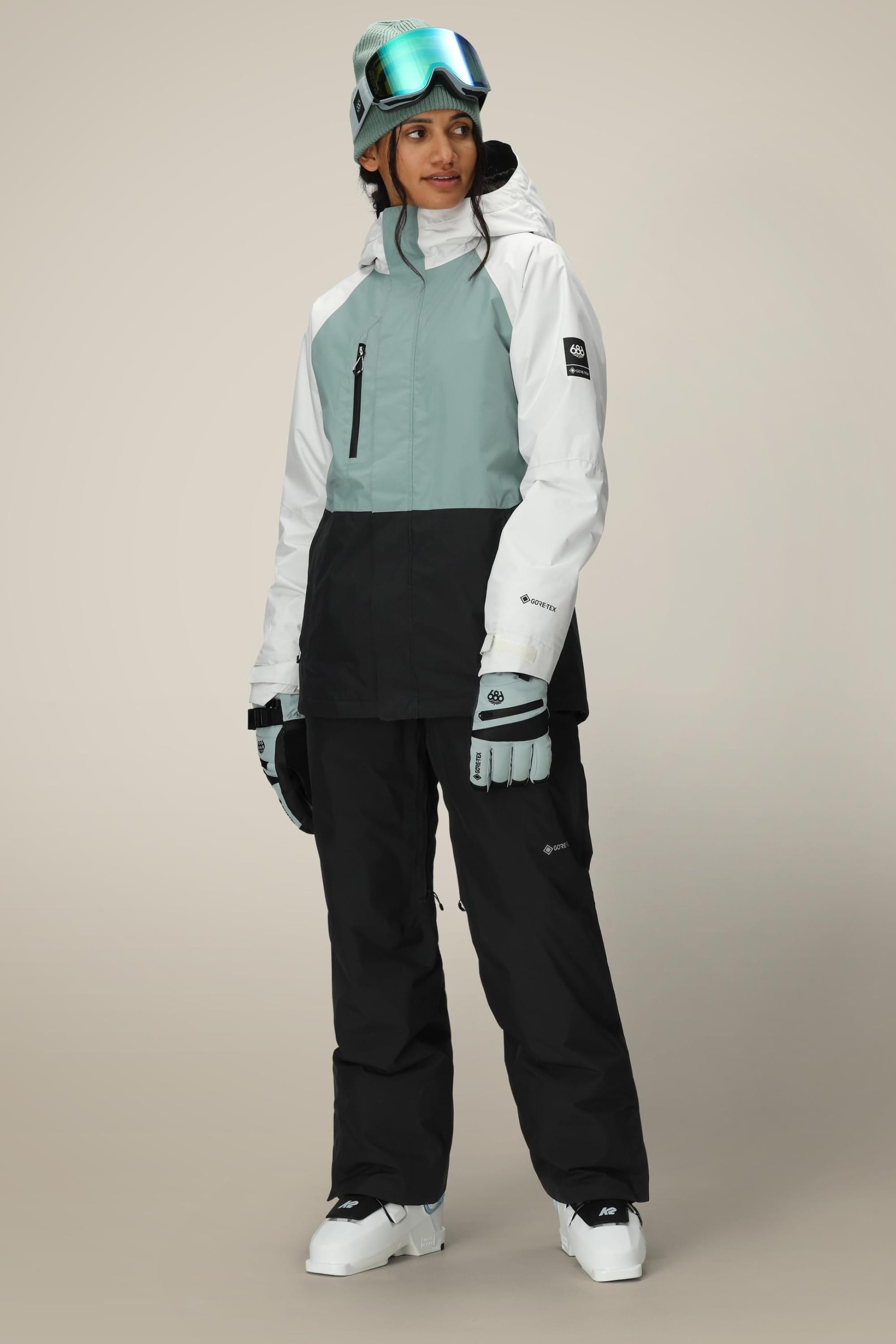 A person wearing a ski helmet, goggles, gloves, and a color-blocked winter jacket with the 686 Women’s GORE-TEX® Willow™ Insulated Pant by 686 and white boots stands facing slightly left against a neutral background.