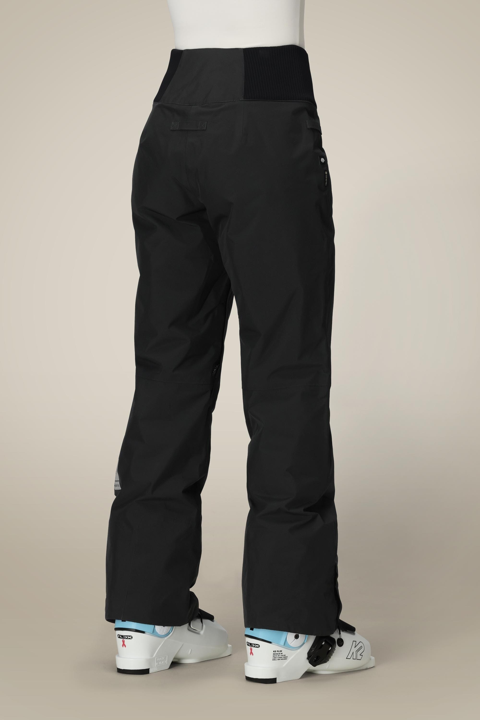 A person is shown from the back wearing 686 Women's GORE-TEX Willow™ Insulated Pant in black with white ski boots, standing against a plain beige background.