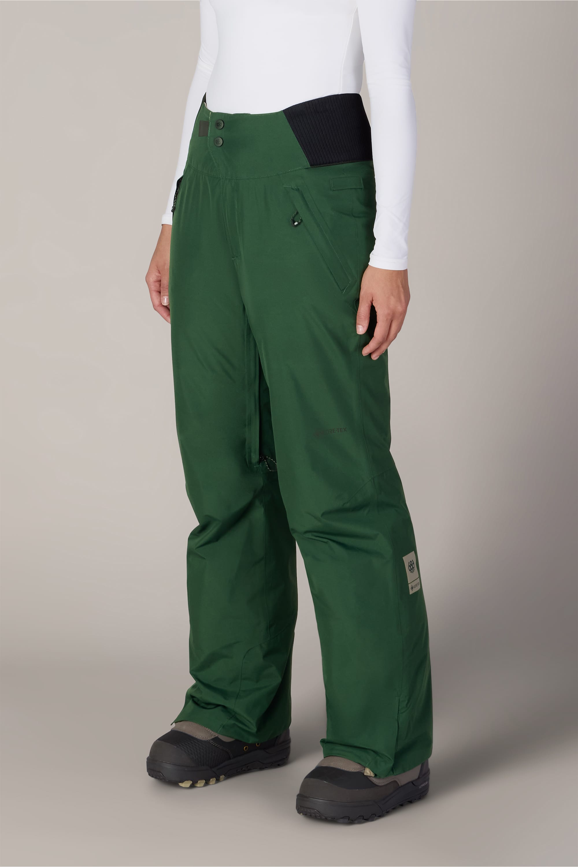 A person is shown from the waist down, wearing the 686 Women’s GORE-TEX® Willow™ Insulated Pant in green and black snow boots with a white long-sleeve top, standing against a plain light background.