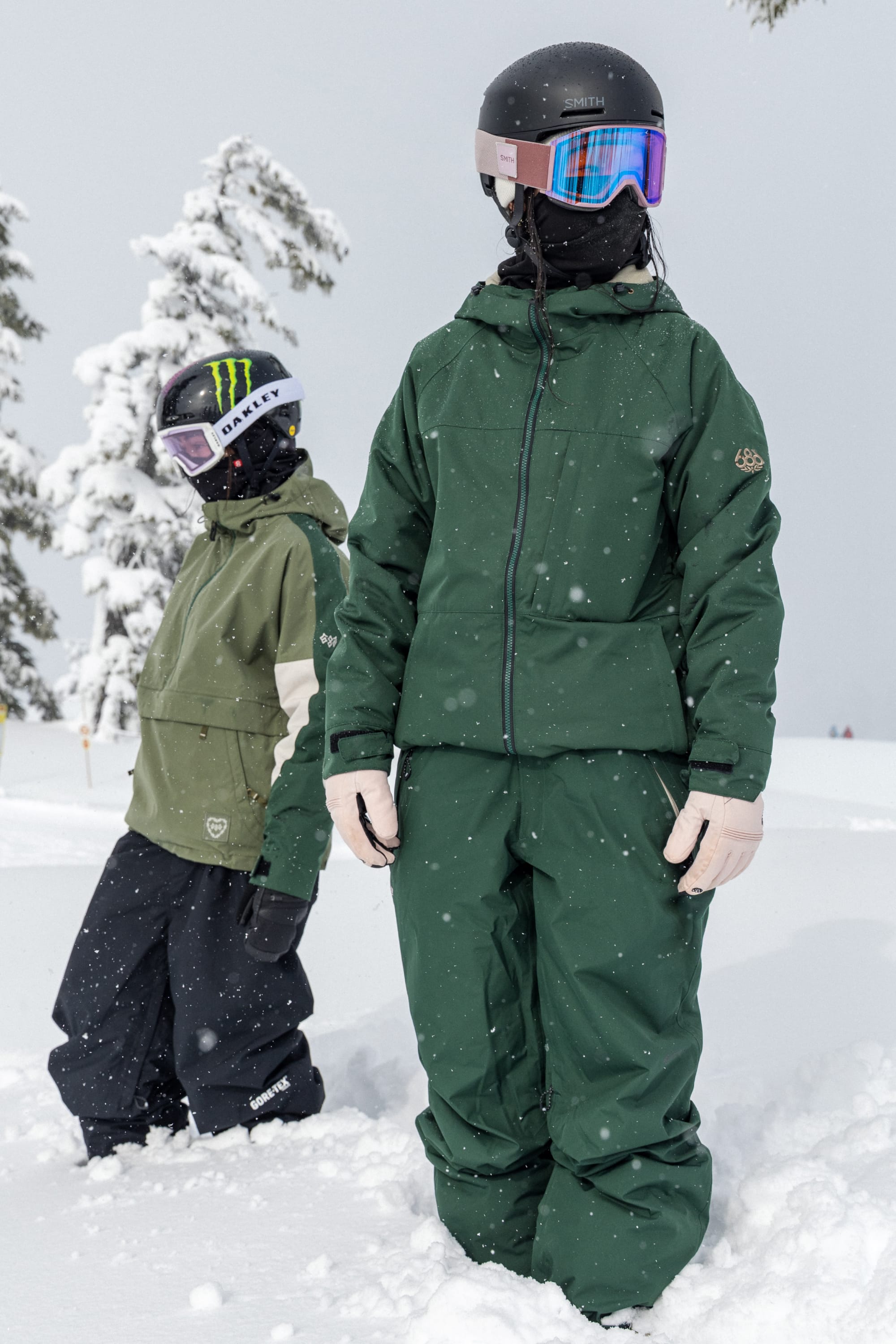 Outfitted in 686 Women’s GORE-TEX® Willow™ Insulated Pants, two people in green winter gear and helmets stand together in the snowy landscape with goggles on, surrounded by snow-covered trees as fresh snow falls on the mountain slope.