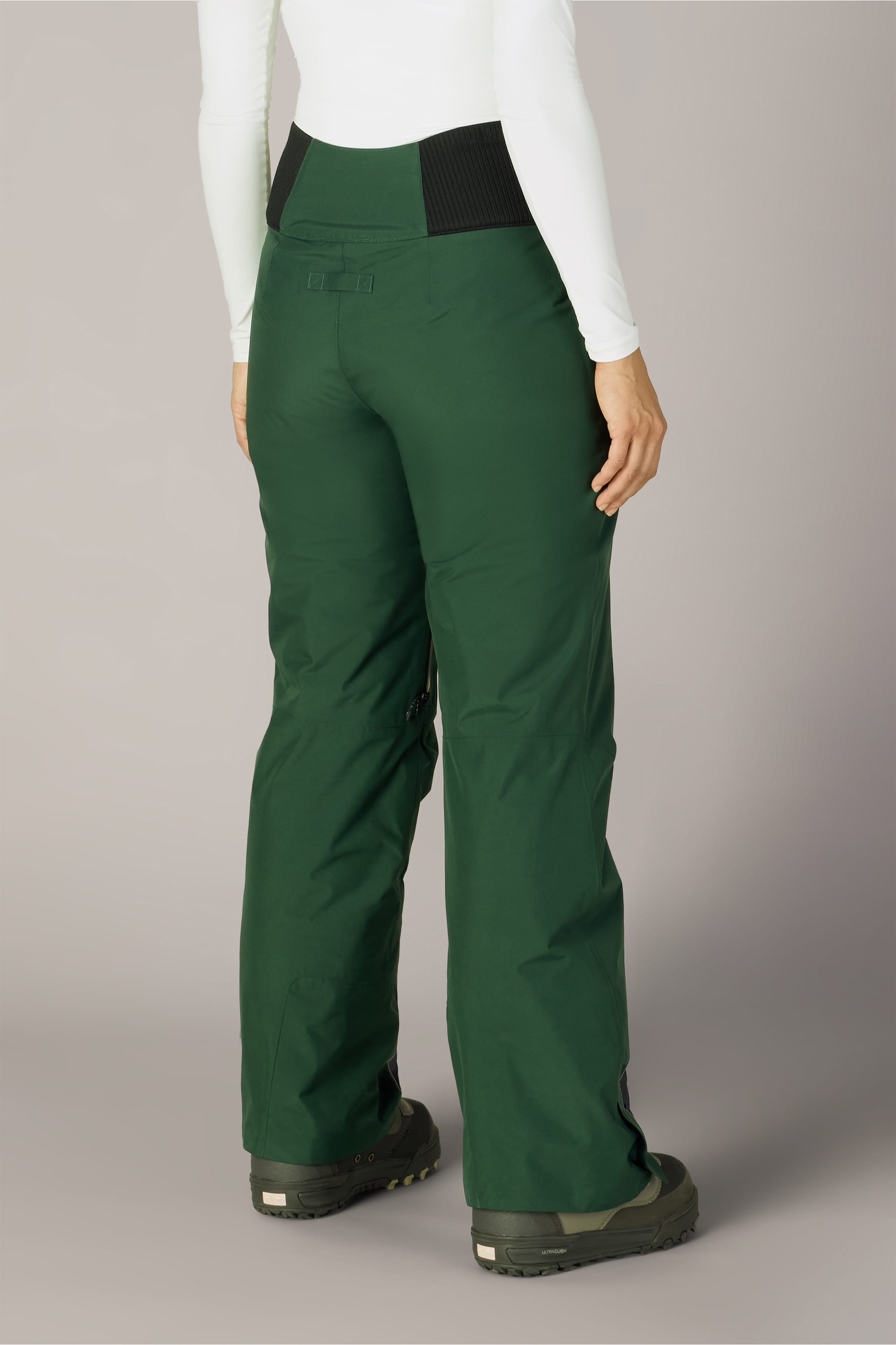 A person is seen from behind, standing against a neutral background, wearing 686 Women’s GORE-TEX® Willow™ Insulated Pant in green, paired with black boots and a long-sleeve white top.