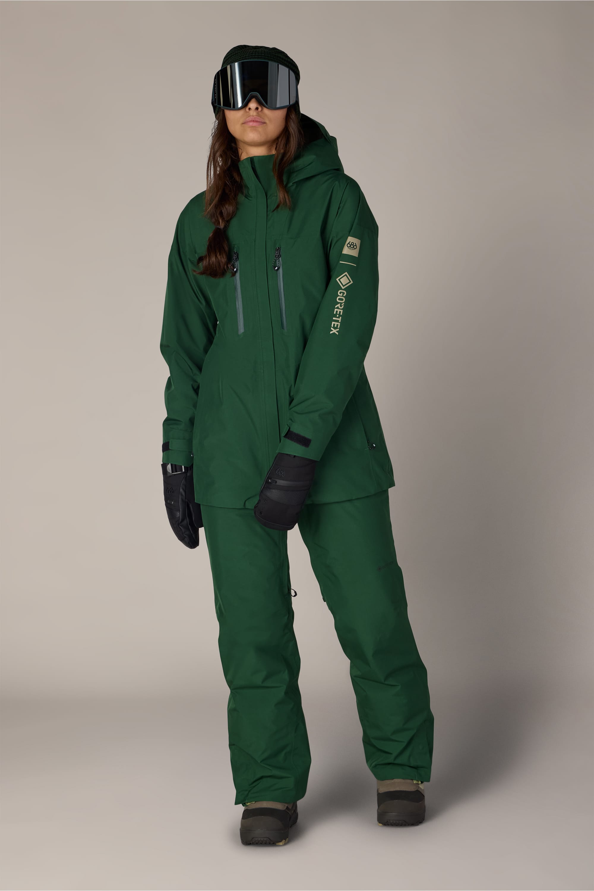 Wearing the 686 Women’s GORE-TEX® Willow™ Insulated Pant, a person stands against a plain light background, dressed in matching green ski attire with black gloves, black snow boots, and large black ski goggles.
