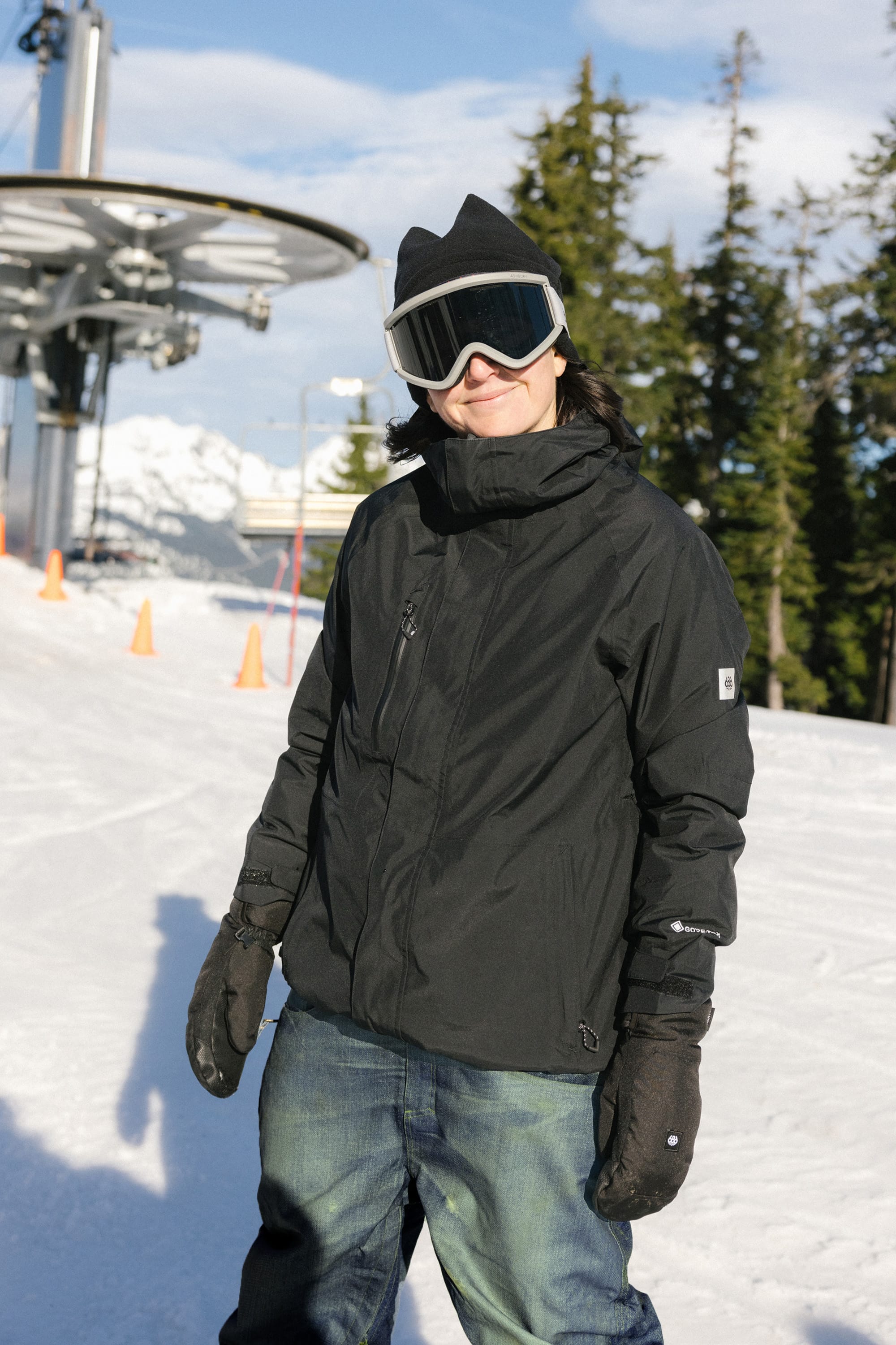 On a sunny snowy slope with trees and ski lifts, a person in a black jacket, gloves, jeans, and ski goggles showcases the 686 Women's Deconstructedª Denim Insulated Pant by 686—stylish waterproof pants ideal for outdoor adventures.