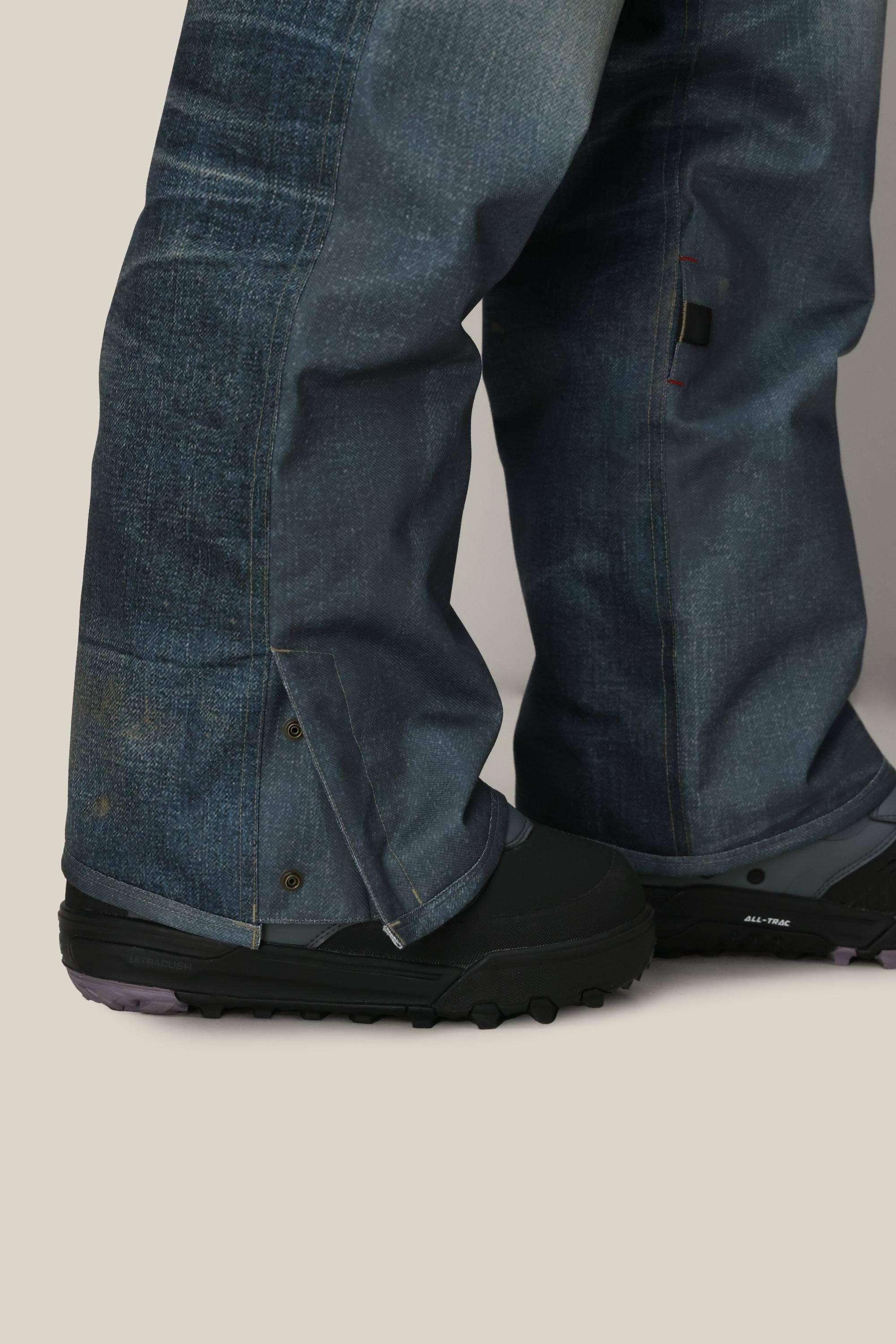 A close-up of a person wearing 686 Women's Deconstructed™ Denim Insulated Pant over black outdoor shoes with rugged soles, standing on a light beige surface. The pants are loose-fitting, faded blue with snap closures and a slightly worn texture.