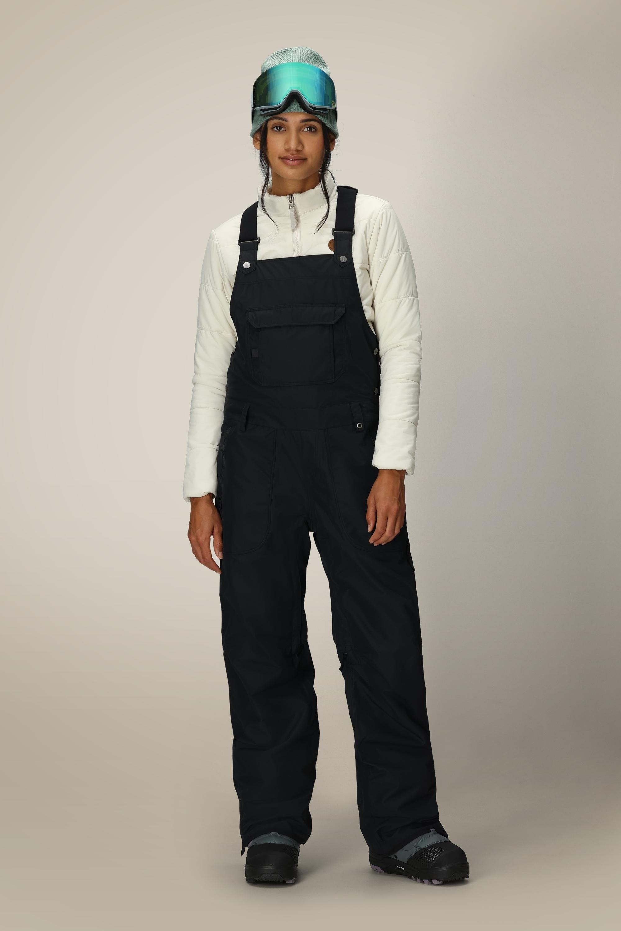 A person wearing a white quilted jacket, 686 Women's Black Magic™ 20K Shell Bib overalls by 686, black boots, a teal helmet, and blue ski goggles stands against a neutral background.