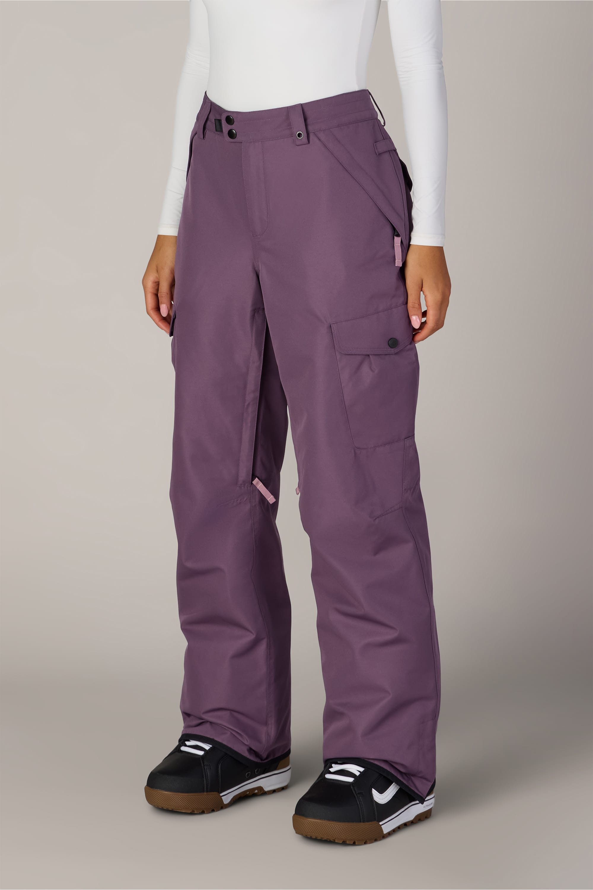 A person wearing the 686 Women’s Aura™ Insulated Cargo Pant in purple, paired with a white long sleeve top and black winter boots featuring white and brown soles, stands against a plain light background.