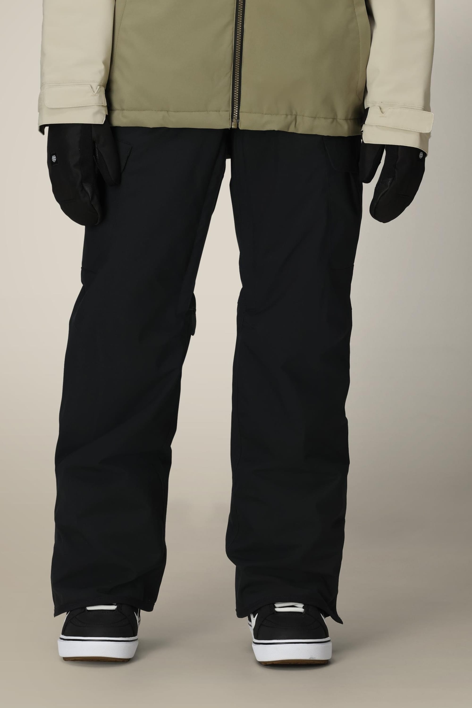 A person wearing 686 Women's Aura™ Insulated Cargo Pant, black gloves, black and white sneakers, and a beige and cream jacket stands against a plain beige background, with only the lower half of their body visible.