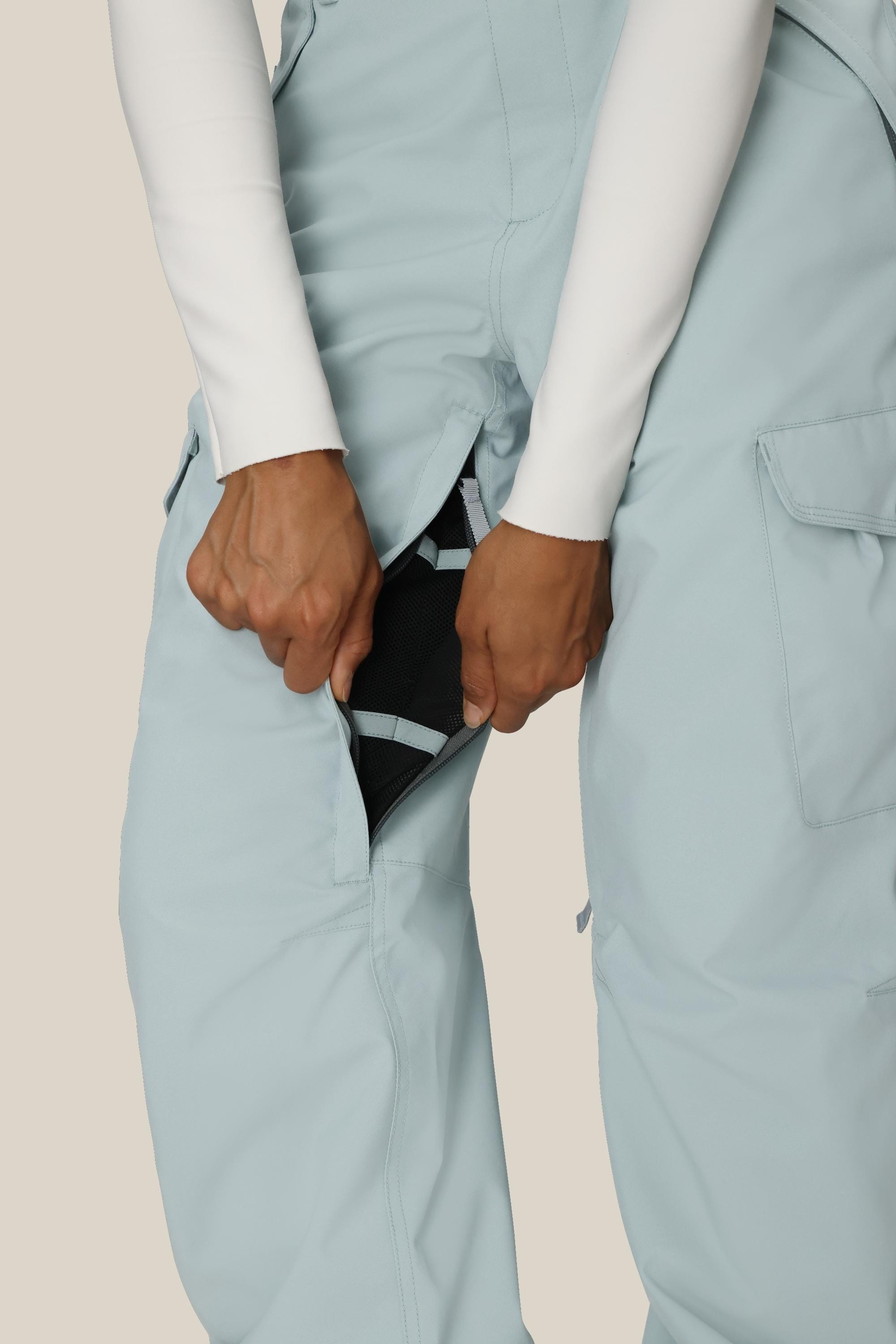 A person in 686 Women's Aura™ Insulated Cargo Pant in light blue and a white long sleeve shirt unzips a side vent, revealing black mesh fabric underneath.