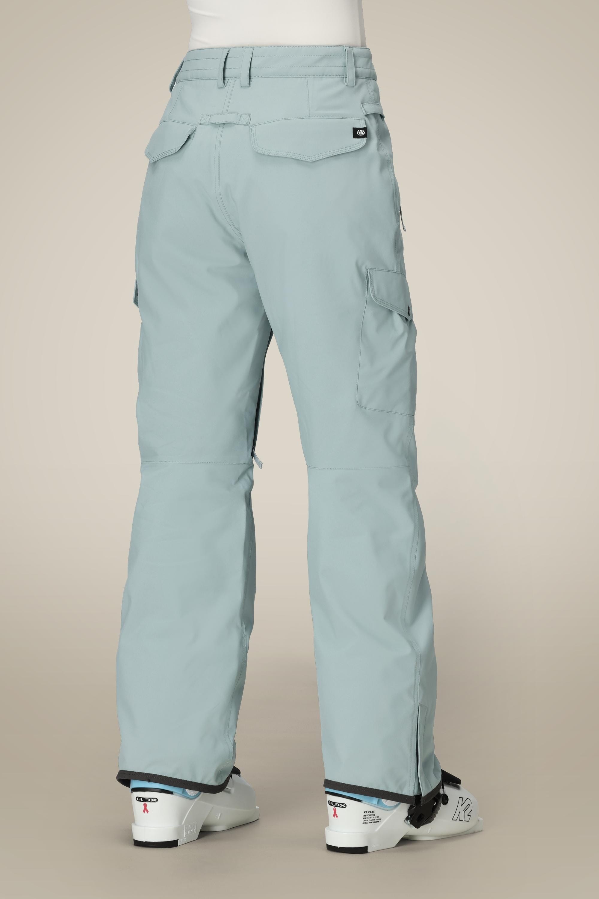 A person wearing light blue 686 Women's Aura™ Insulated Cargo Pants with flap pockets and a relaxed fit, along with white ski boots, stands with their back to the camera against a plain beige background.