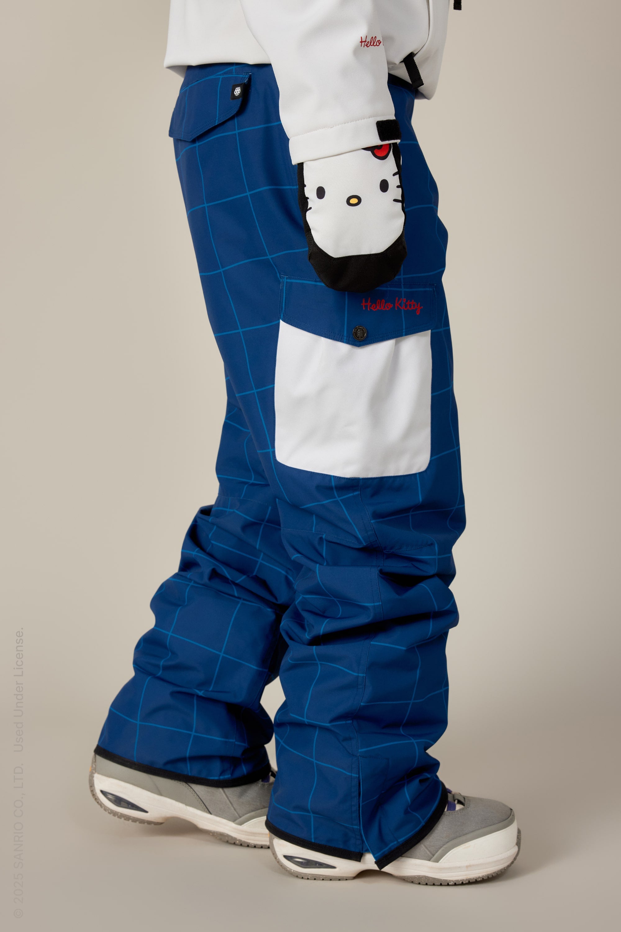 A person wears 686 Women's Aura™ Insulated Cargo Pant in a blue checkered design with a white pocket, paired with white and black Hello Kitty gloves, a white jacket, and gray winter boots, standing against a neutral background.