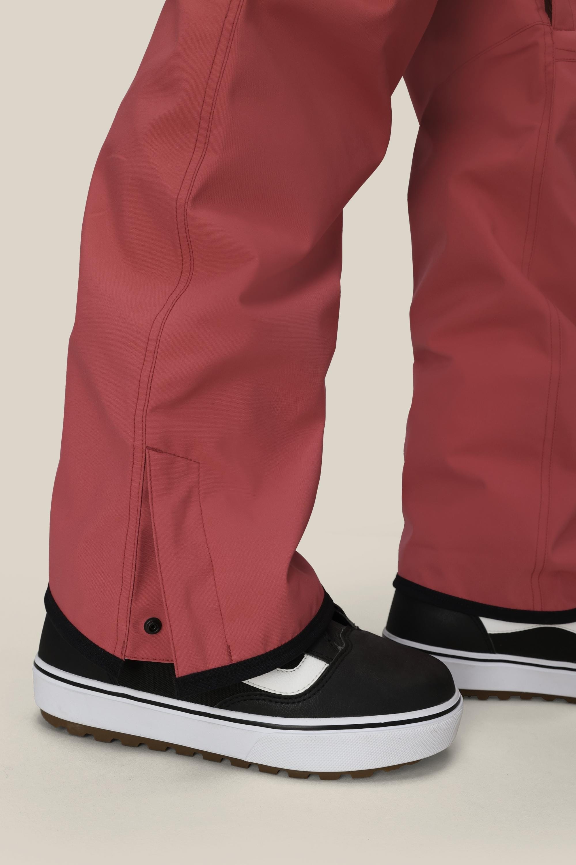 Close-up of a person wearing the 686 Women's Aura™ Insulated Cargo Pant in a loose fit, rose color, paired with black-and-white sneakers. The pants feature snap button detail near the ankle and slightly cover the shoes, set against a plain light background.
