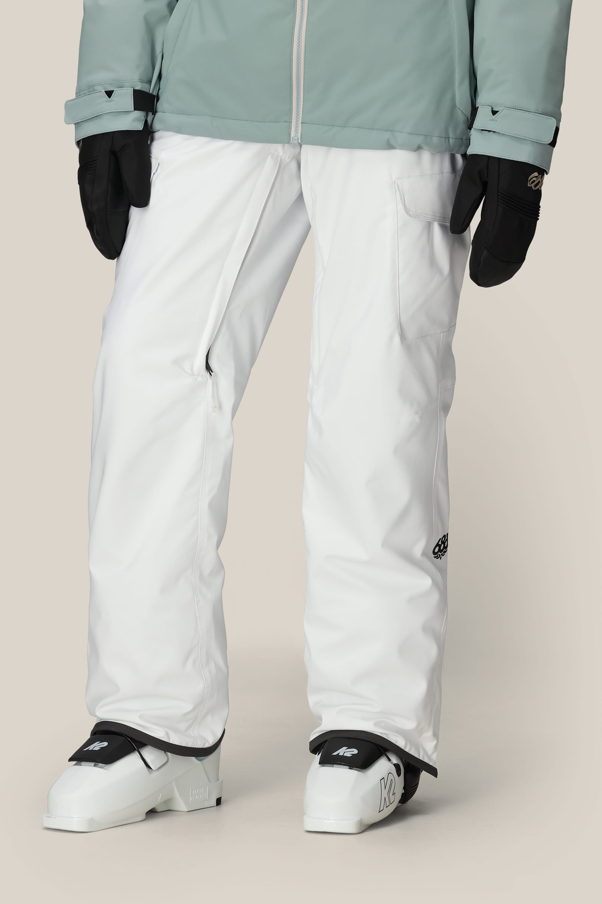 A person wearing 686 Women's Aura™ Insulated Cargo Pant, black gloves, a light green jacket, and white ski boots stands against a neutral background.