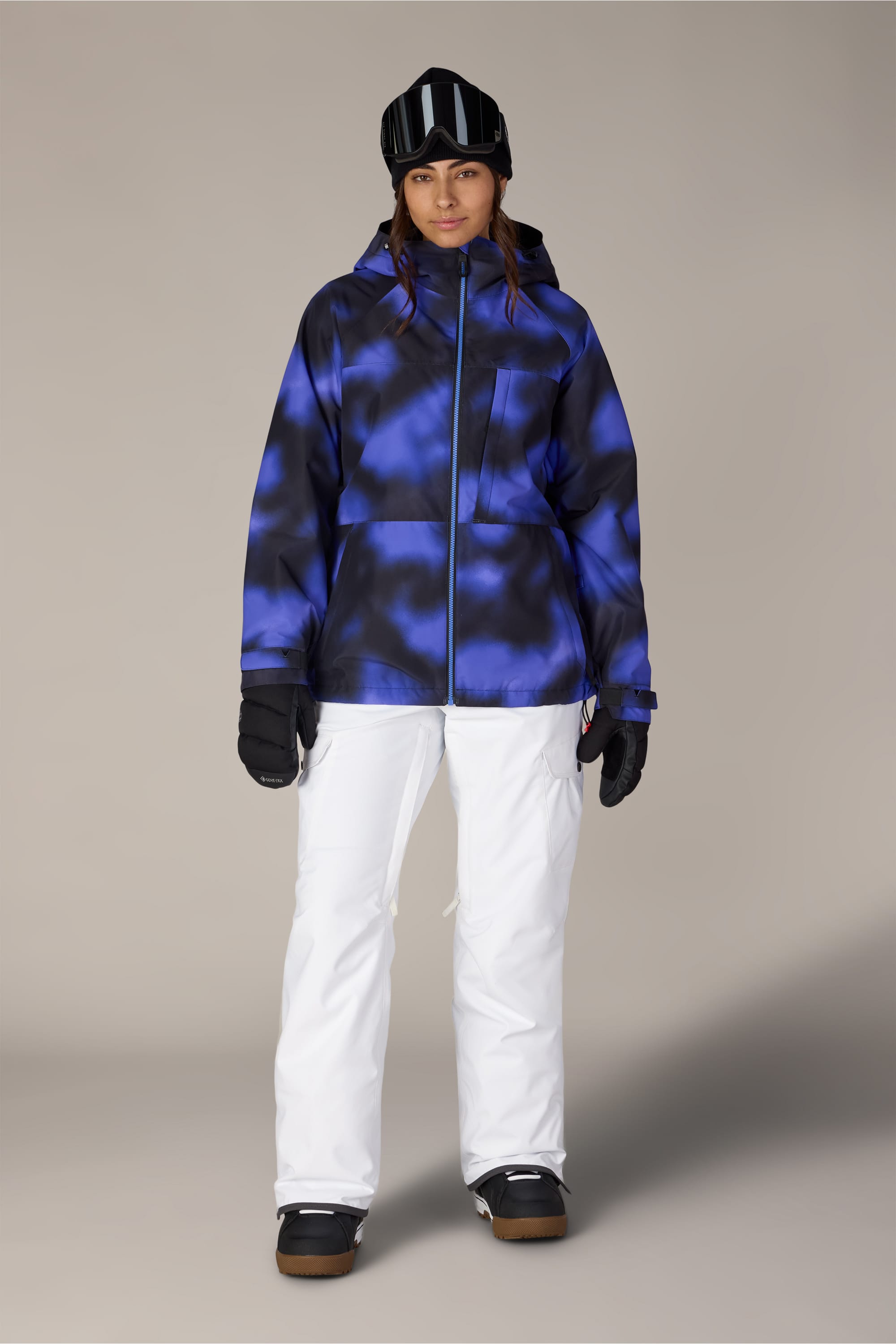 A person stands facing forward, wearing a blue and black tie-dye winter jacket, black gloves, black goggles, and a black beanie, paired with the 686 Women’s Aura™ Insulated Cargo Pant by 686 in white—ready for snow sports against a plain background.