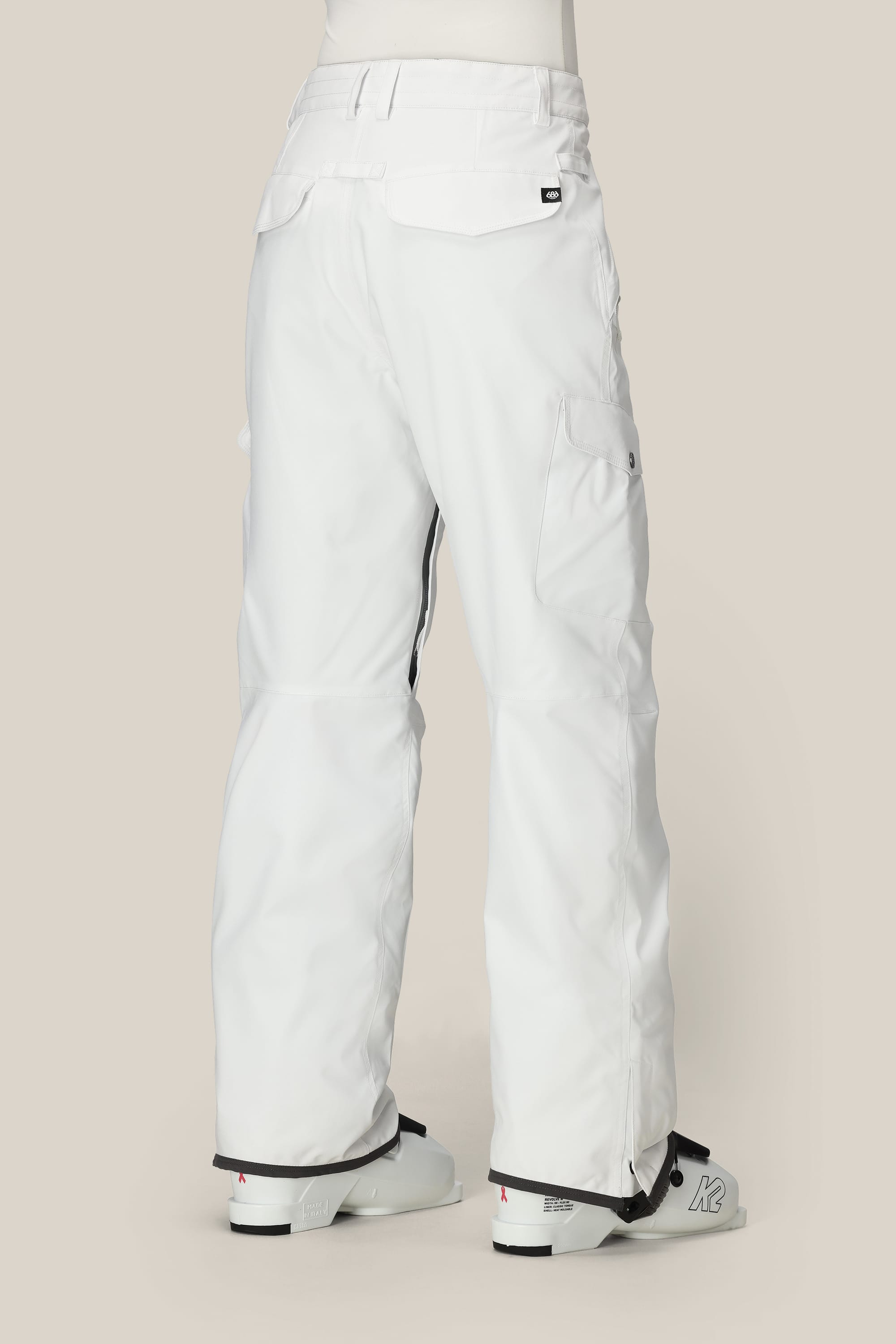 A person is shown from the back wearing 686 Women's Aura™ Insulated Cargo Pant in white, featuring pockets and a relaxed fit, along with white ski boots, against a plain light beige background.