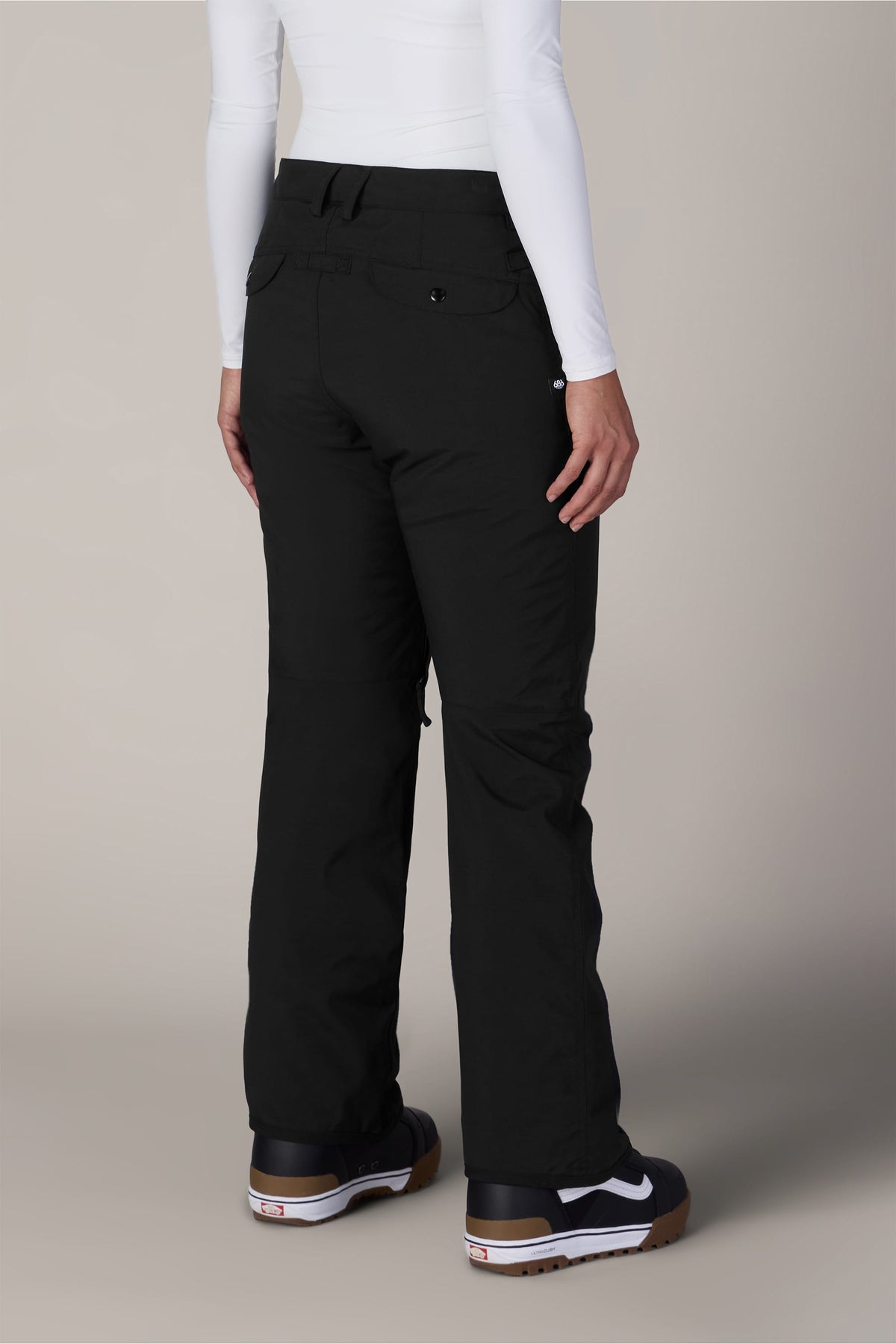 A person wearing the 686 Women’s Standard Shell Pant by 686, paired with a white long-sleeve shirt and black sneakers with white soles, is standing with their back to the camera against a plain background.