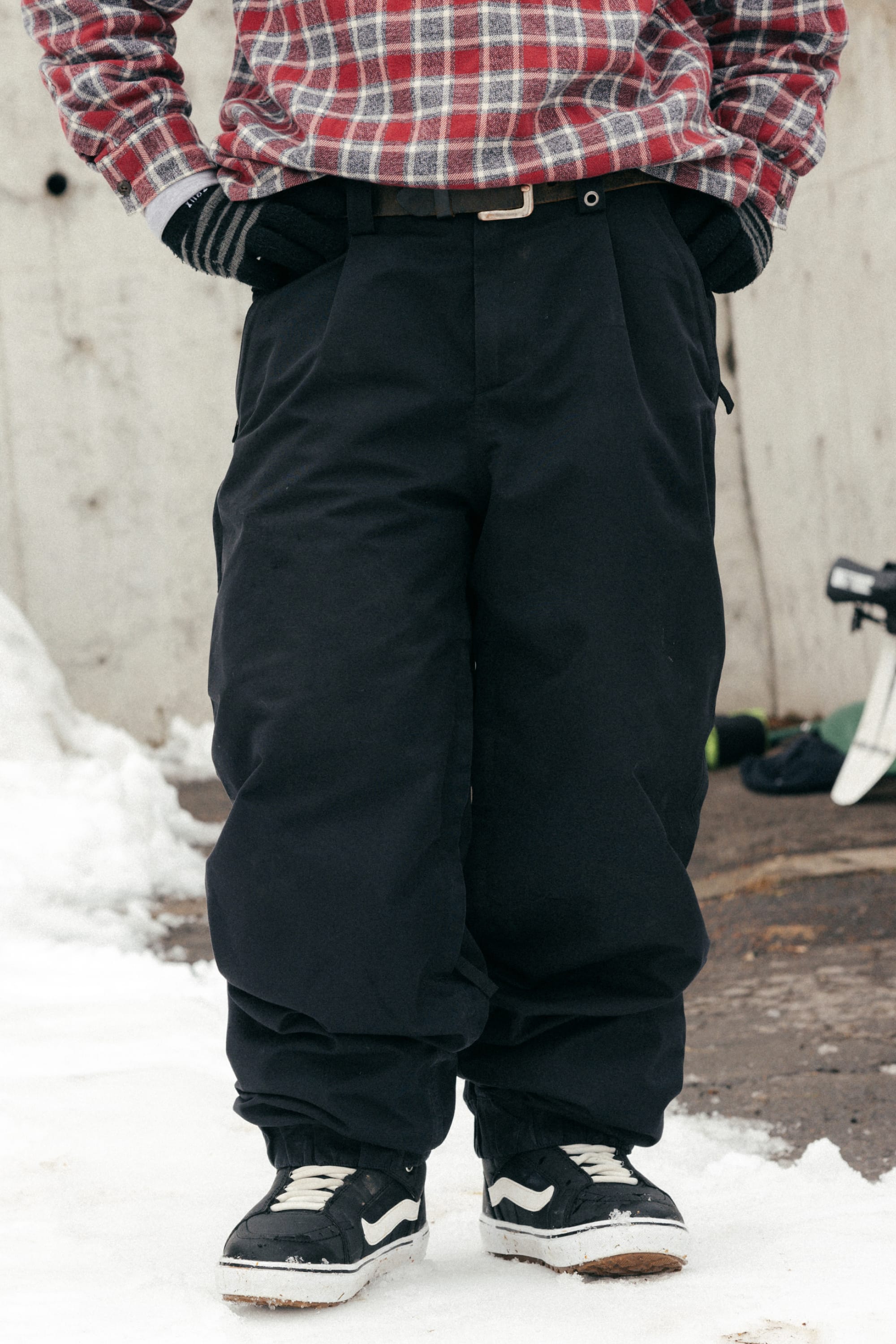 A person stands on snow wearing 686 Women's Outline™ Shell Pants by 686, black gloves, black-and-white sneakers, and a red plaid shirt with hands in pockets. A snowboard lies nearby.
