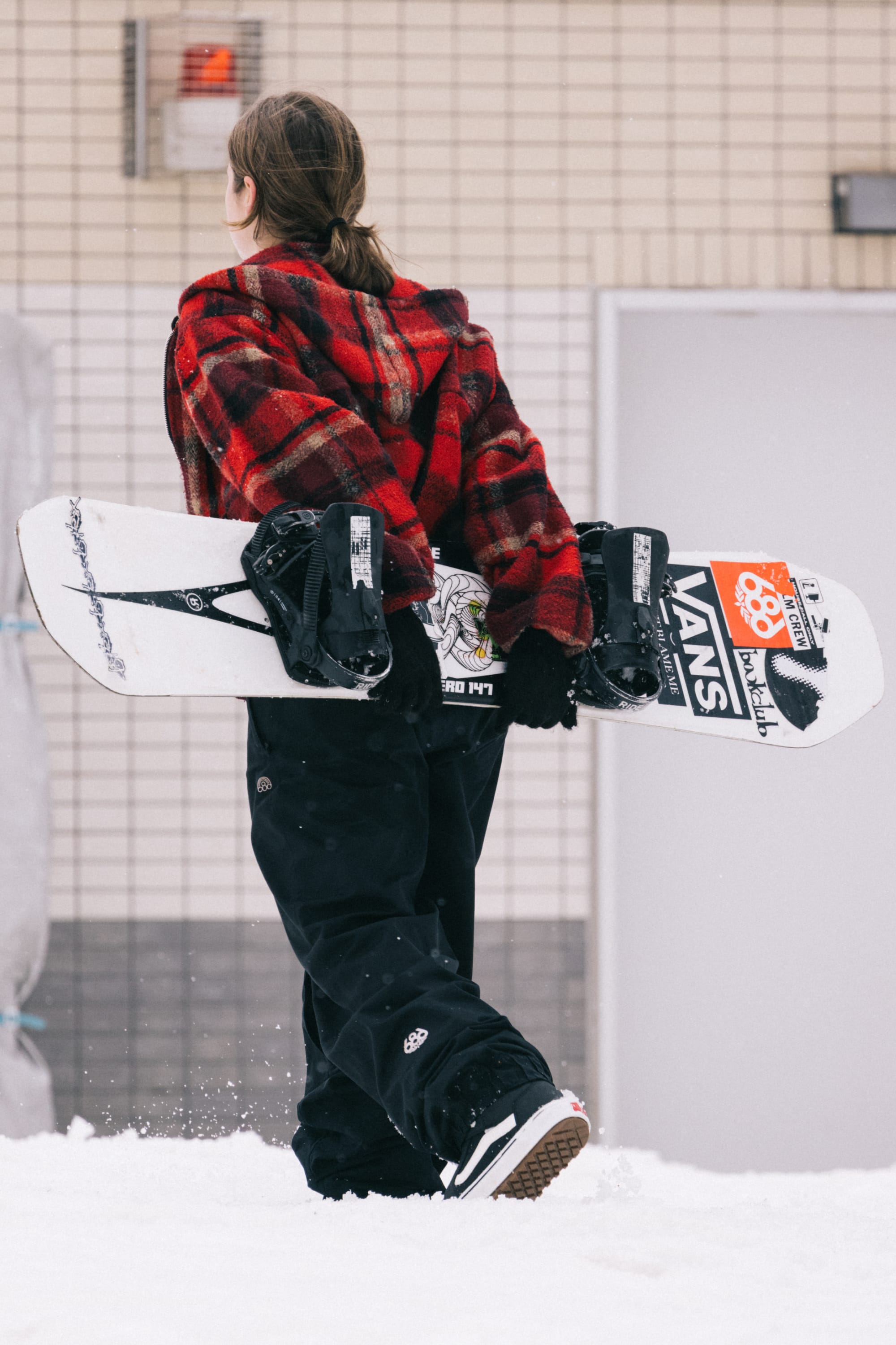 Wearing the 686 Women's Outline™ Shell Pant, a person in a red plaid jacket walks on snow with a snowboard featuring VANS stickers, set against a tiled wall and door.