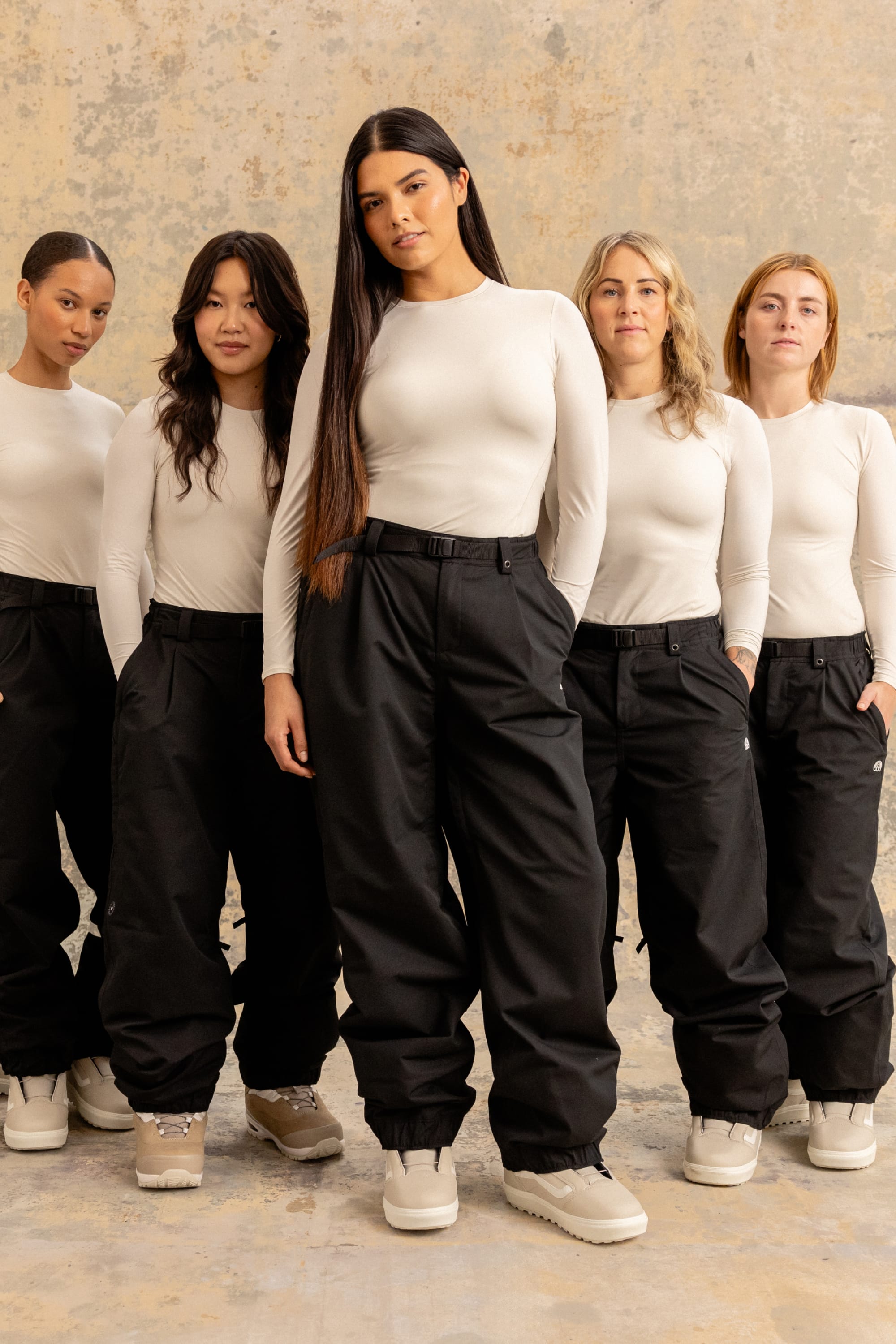 Five women stand together against a textured backdrop, all wearing matching 686 Women's Outline™ Shell Pants with drawstring cuffs and white long-sleeve tops. Their expressions are serious, and they each have different hairstyles.