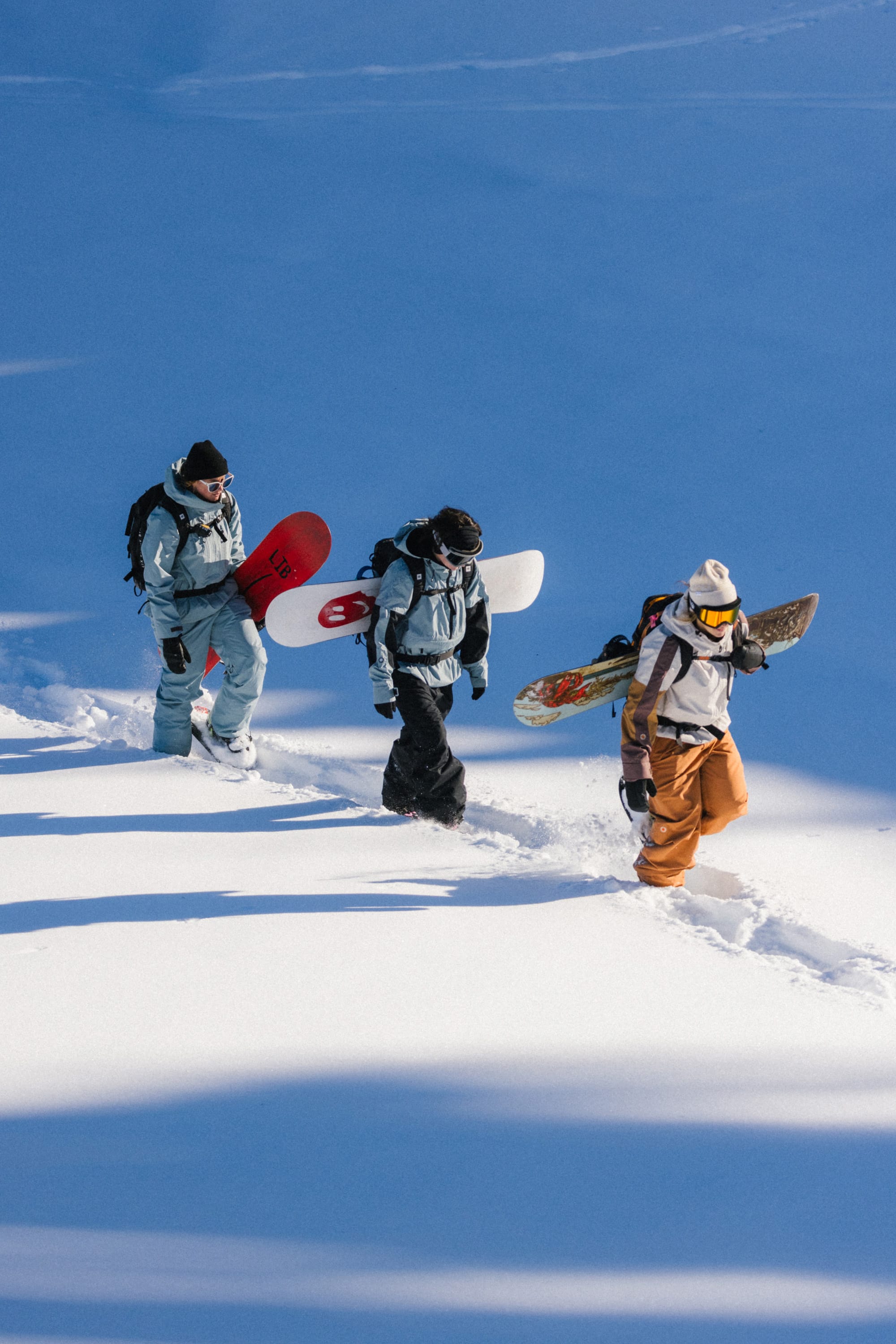 Three snowboarders in winter gear, featuring the 686 Women's Outline™ Shell Pant by 686, walk single file through deep snow, carrying their boards on their backs across a snowy landscape under bright sunlight.