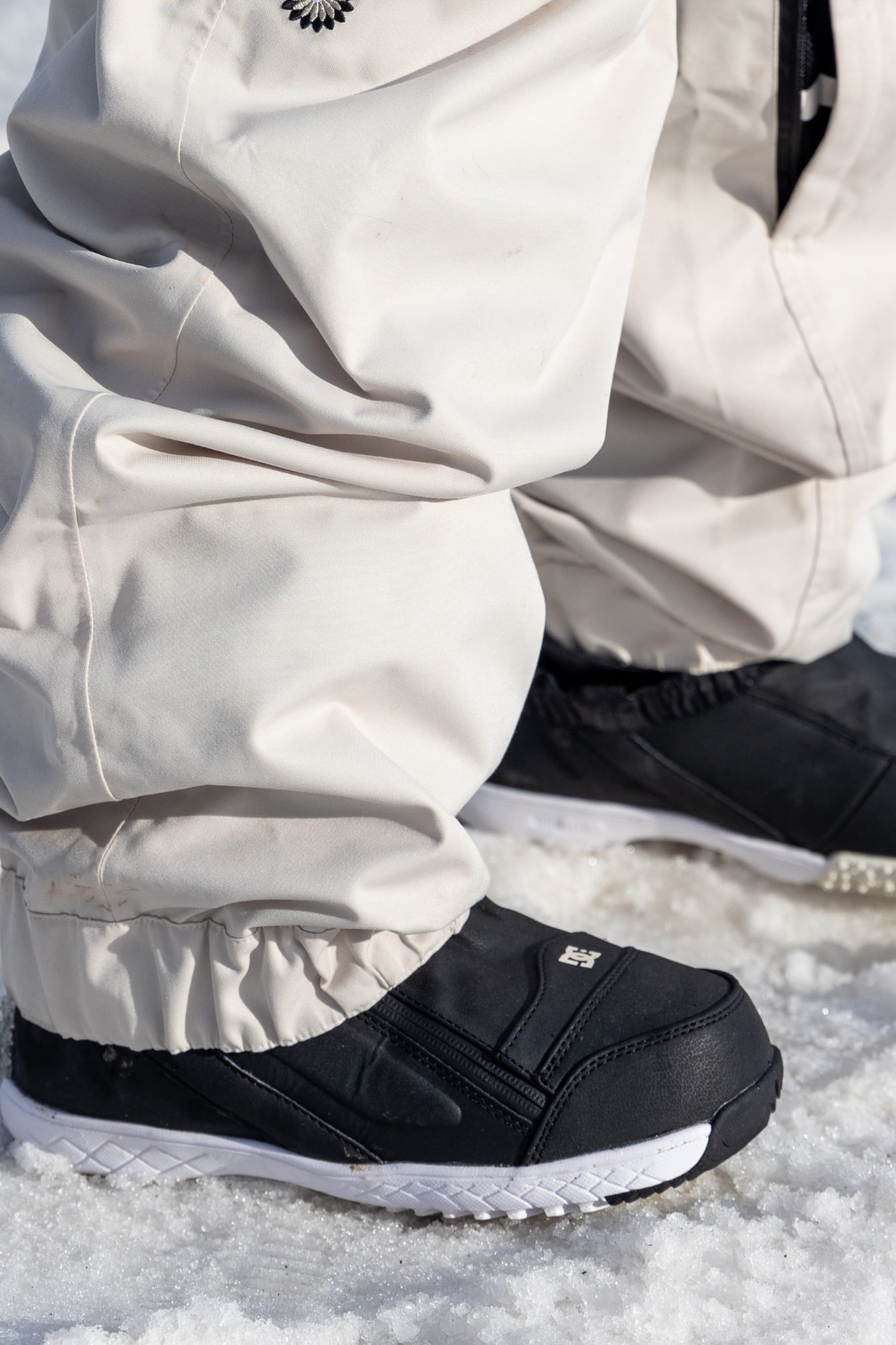 Close-up of a person wearing 686 Women's Outline™ Shell Pant in beige with black winter shoes, standing on snow. The image focuses on the lower legs and shoes.
