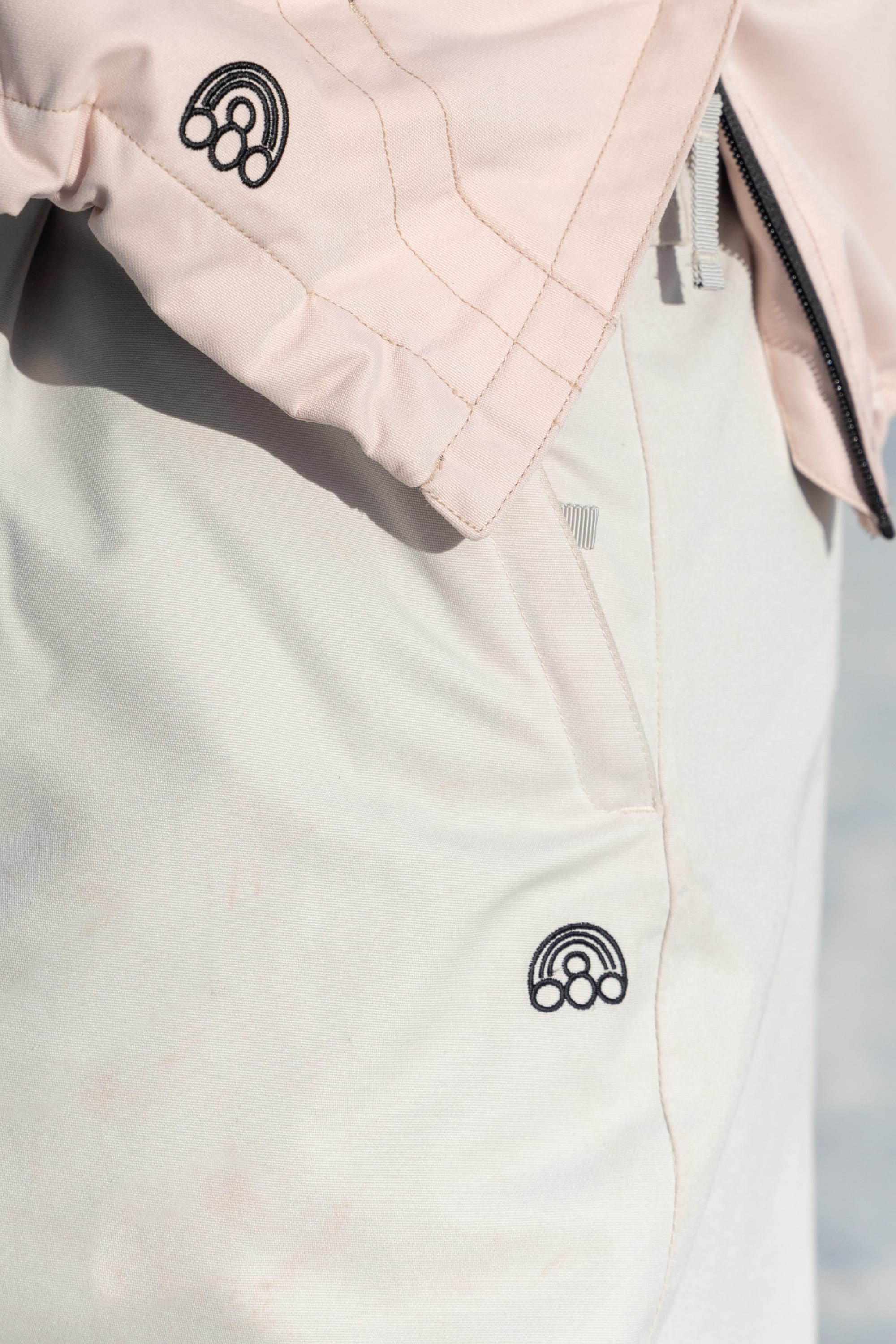 Close-up of light pink jacket and beige 686 Women's Outline™ Shell Pant, both featuring a small black embroidered rainbow logo near the pockets.