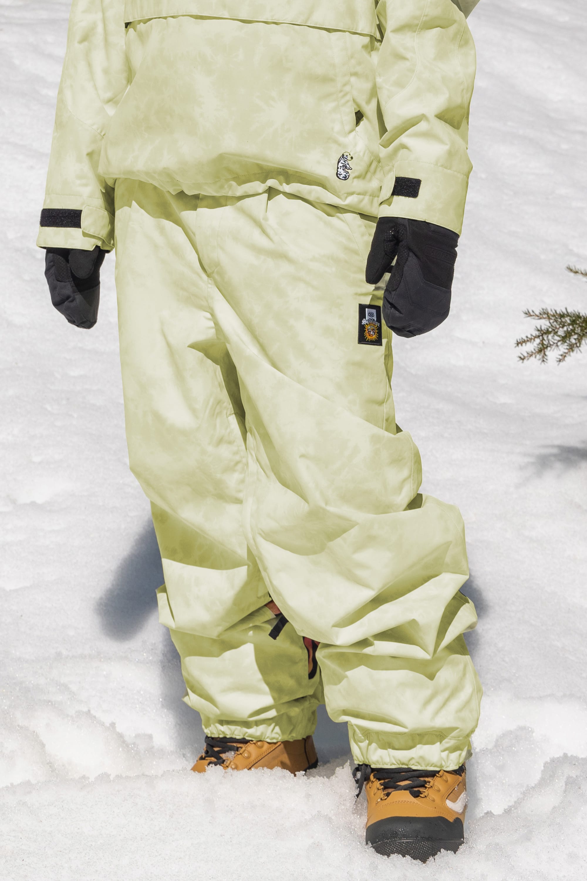 A person wearing 686 Womens Sublime Shell Pant in pale green stands on snow, paired with a matching jacket, black gloves, and tan snow boots. Only the lower half of their body is visible.