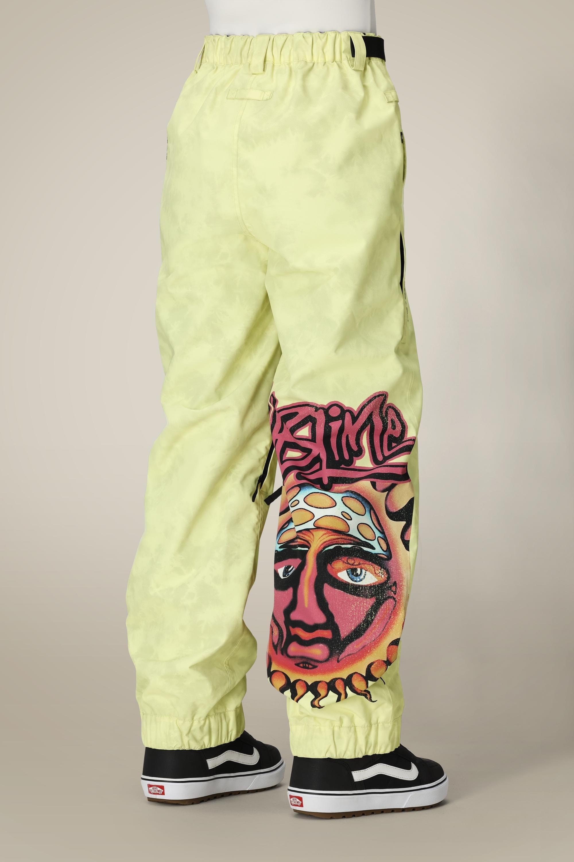 A person wearing 686 Womens Sublime Shell Pant in light yellow with a sun face and SLIME graffiti on one leg, paired with black and white Vans sneakers, shown from the back.