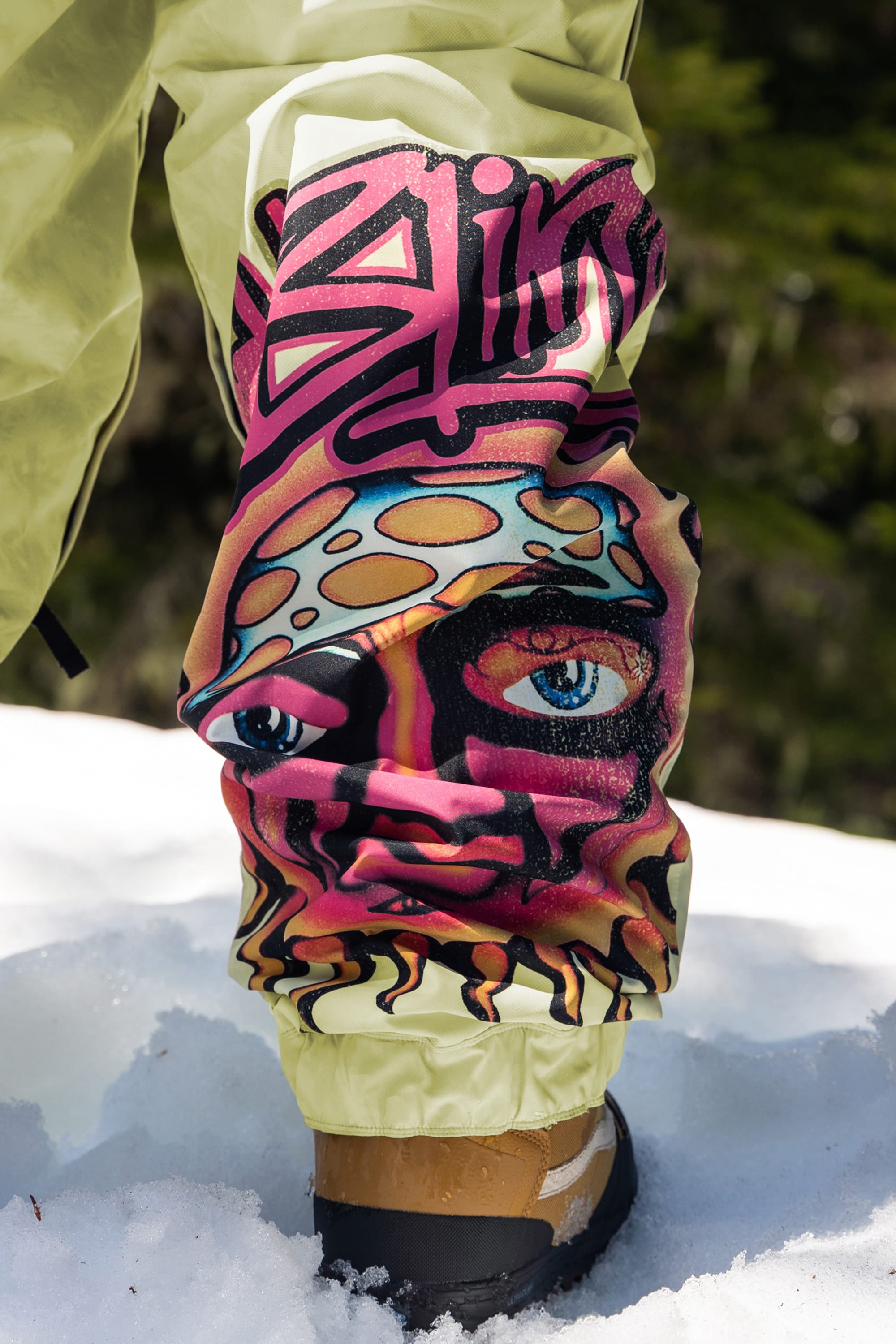 A close-up of the 686 Womens Sublime Shell Pant, featuring graffiti-style art with a blue-eyed face and mushroom hat, stands on snow with blurred trees in the background.