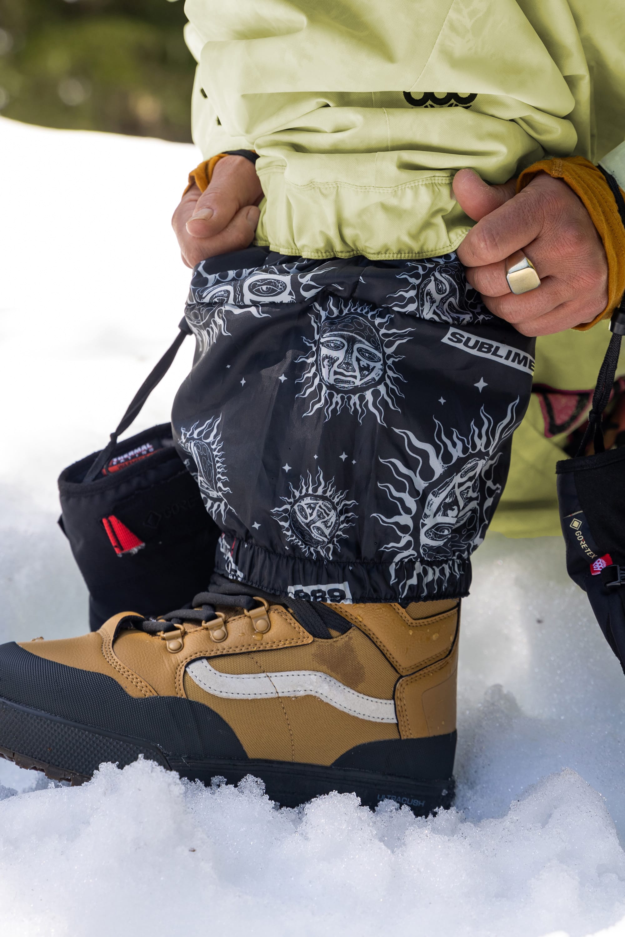 Wearing 686 Womens Sublime Shell Pant, a person in tan and black Vans boots adjusts black snow gaiters with sun and face patterns and Sublime text, standing on snow.
