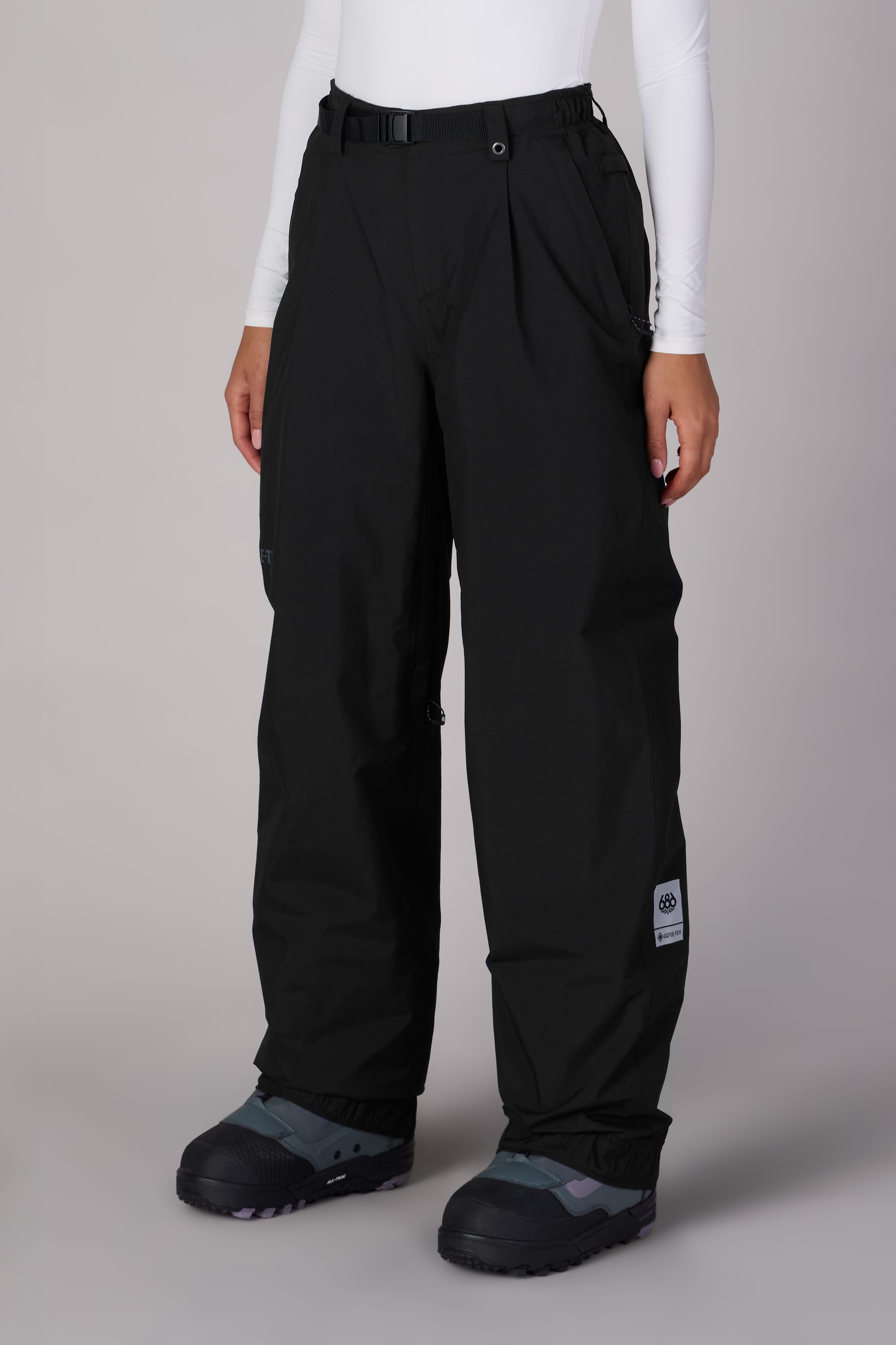 A person wears 686 Women's GORE-TEX Outline™ Shell Pant by 686—loose-fitting black waterproof pants with a belt and ankle patch—paired with a white long-sleeve top and black snow boots, standing on a neutral background.