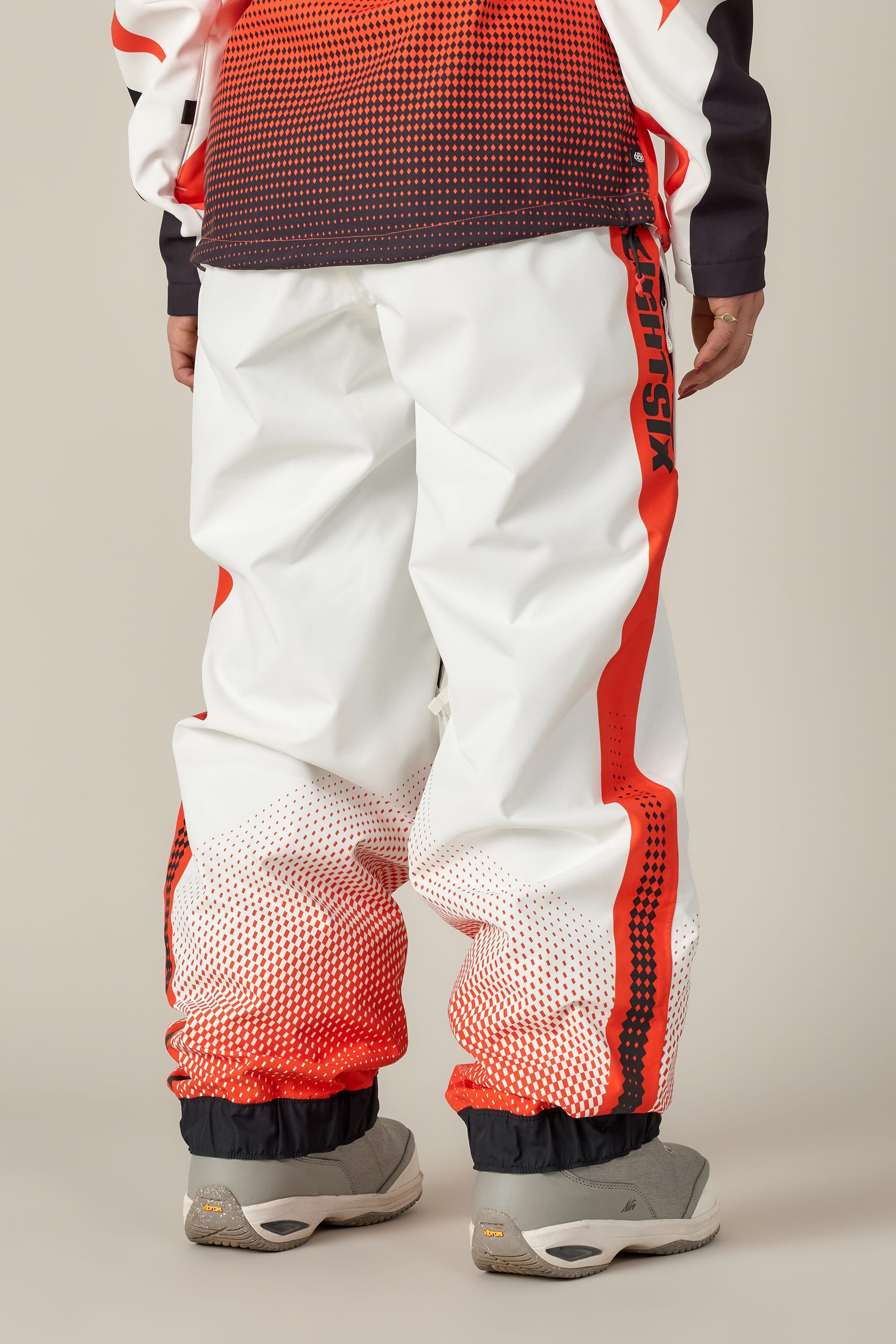 A person is seen from behind wearing 686 Women's Fox Racing Shell Pant by 686, featuring white fabric with red gradient dots and "ANTISOCIAL" on the side, paired with light gray sneakers against a plain background.