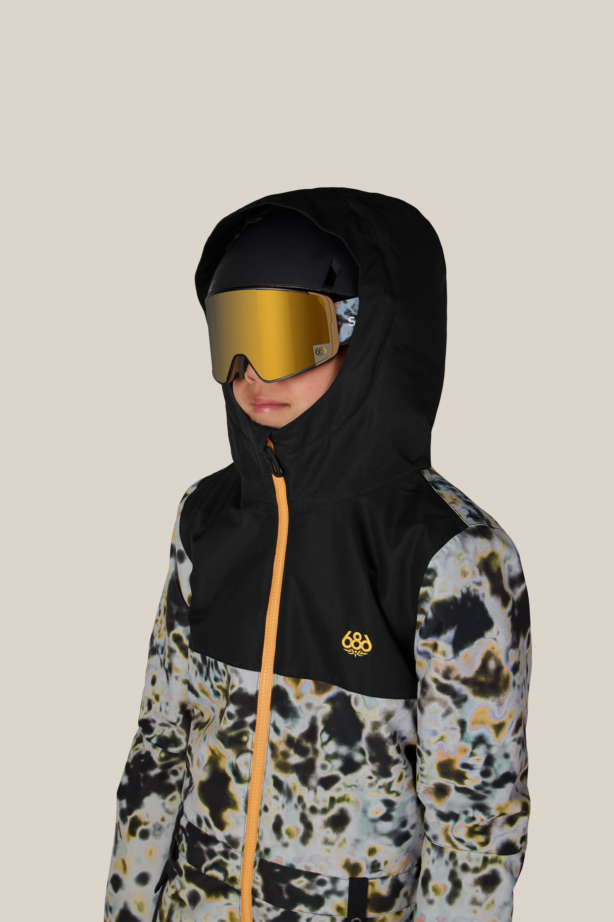 A child wearing the 686 Boys’ Shazam Insulated One-Piece by 686, featuring a black hooded design with a yellow zipper and abstract pattern, paired with large reflective ski goggles and a helmet, stands against a plain beige background.