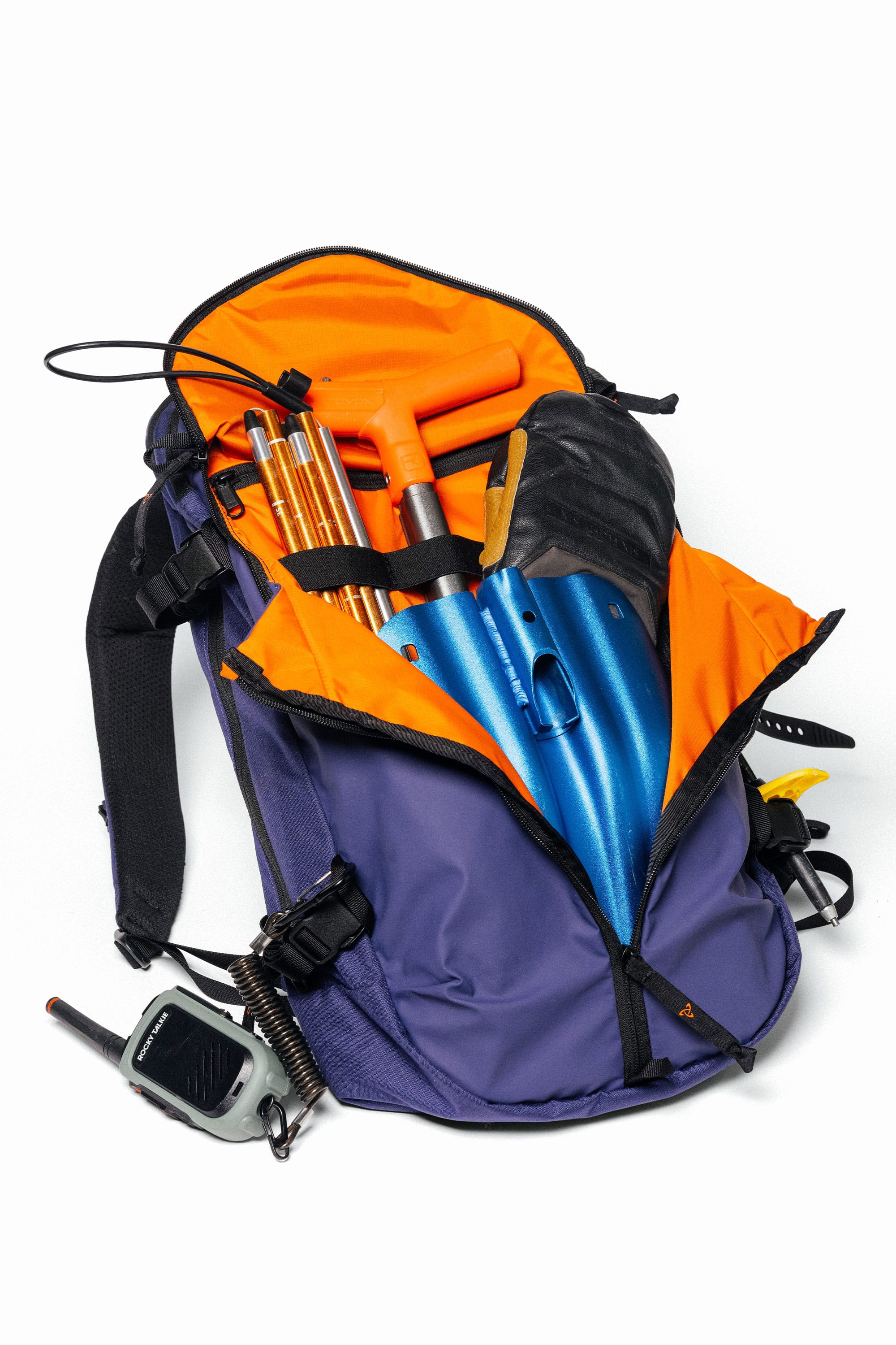 The 686 Mystery Ranch x 686 Qi 22L Snow Pack is unzipped to reveal avalanche safety gear—a collapsible shovel, probe, and walkie-talkie attached to the shoulder strap—displayed on a white background.
