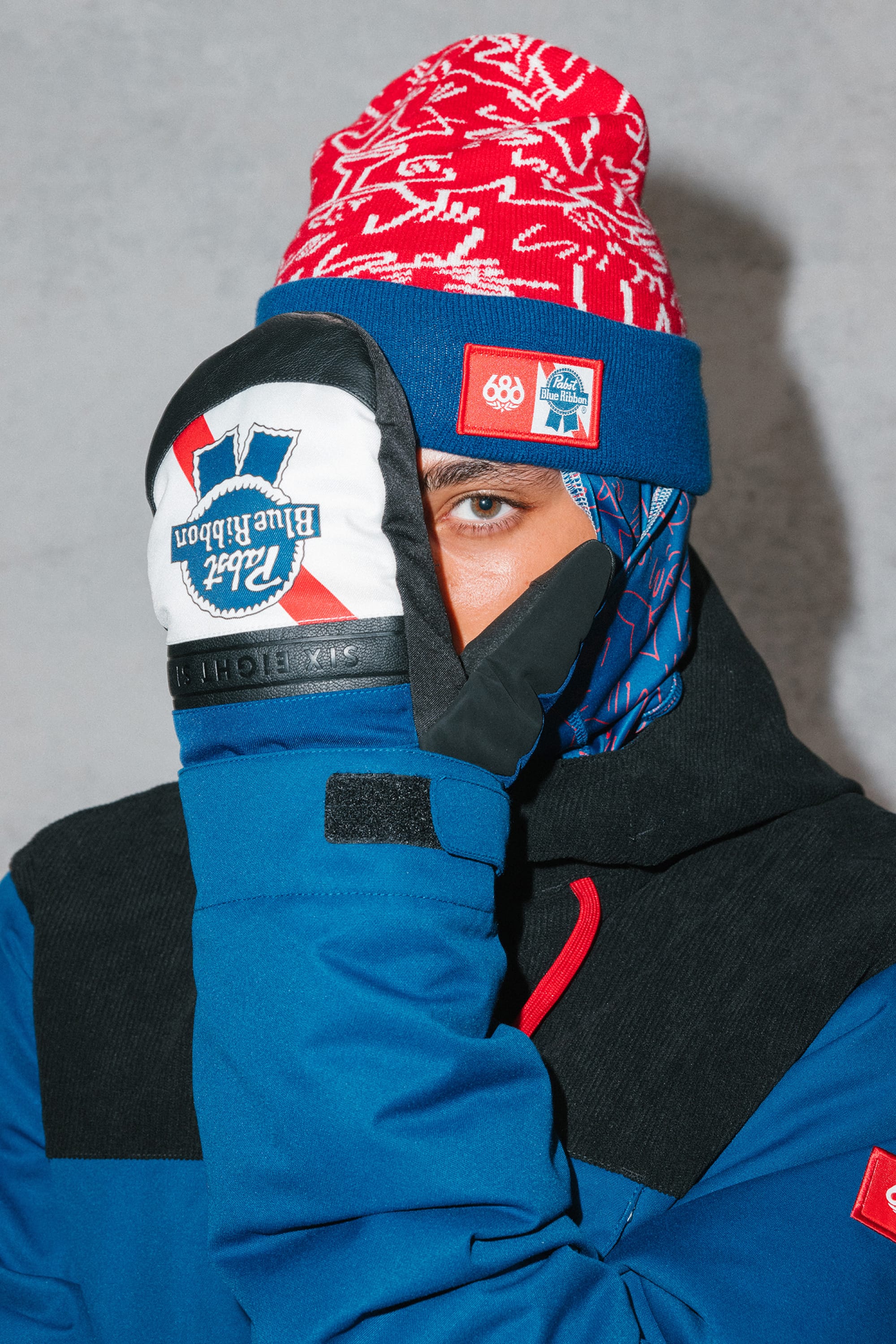 A person in a 686 PBR Beanie, blue jacket, and white Blue Ribbon mittens covers half their face with a gloved hand while looking directly at the camera against a plain gray background.