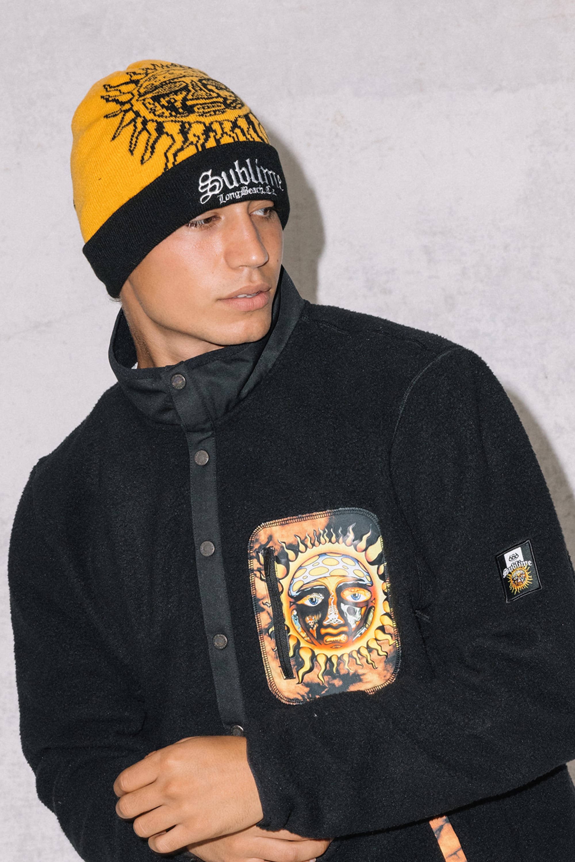 A person wears the 686 Sublime Beanie—a yellow acrylic knit hat by 686—paired with a black jacket featuring a sun design on the pocket, standing against a light grey textured background.
