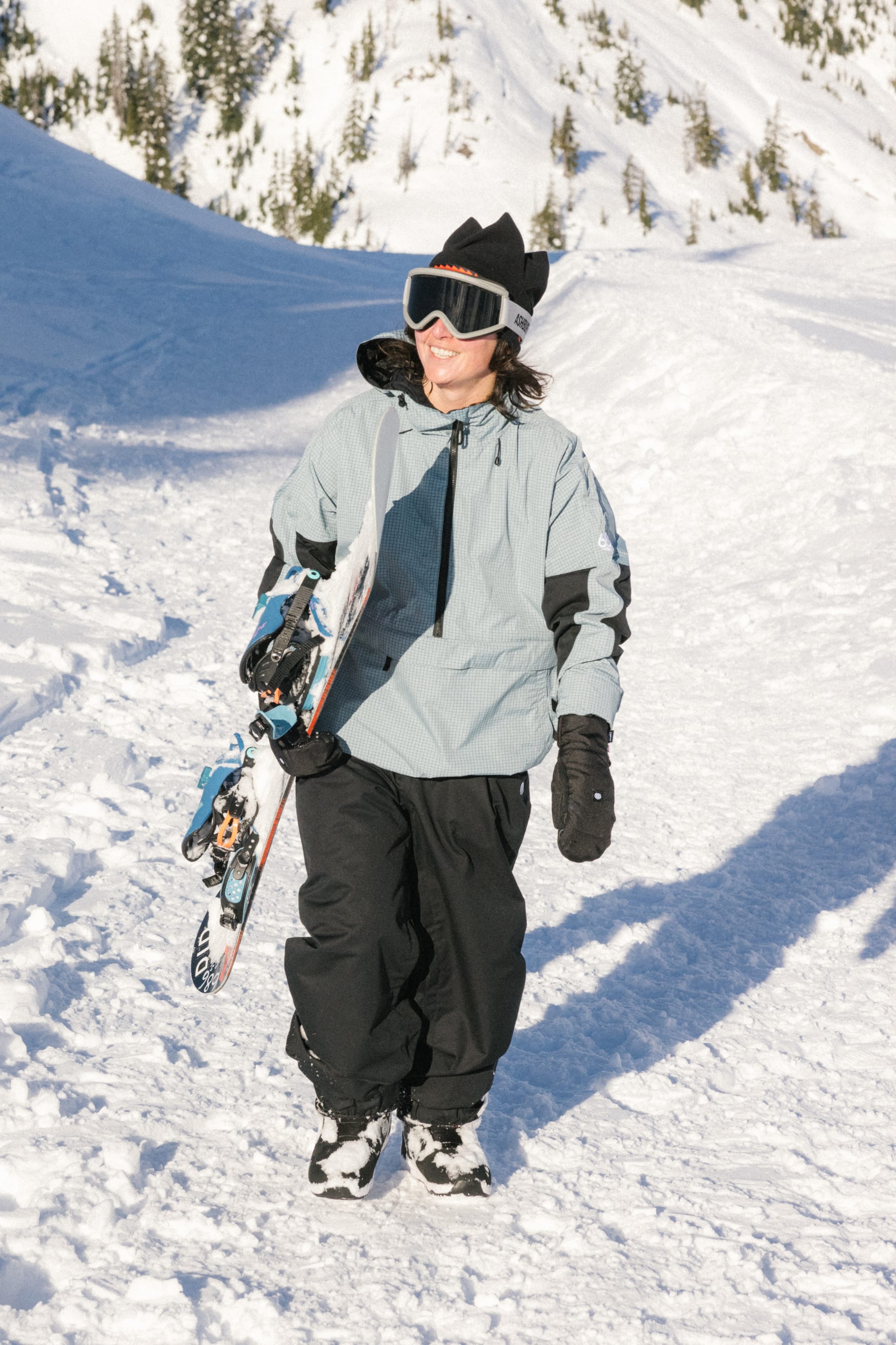 Wearing the 686 4 Points Fleece Hat by 686, a person in winter gear smiles while carrying skis over one shoulder, walking on a snowy mountain slope with trees in the background.