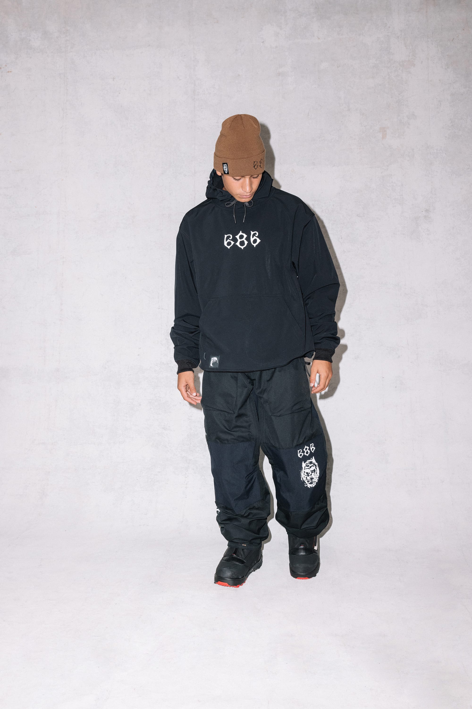 A person wearing the 686 Cooper Beanie, a black hoodie with 888 on the front, black pants with a white tiger graphic, and black shoes stands against a plain light gray background and looks down.