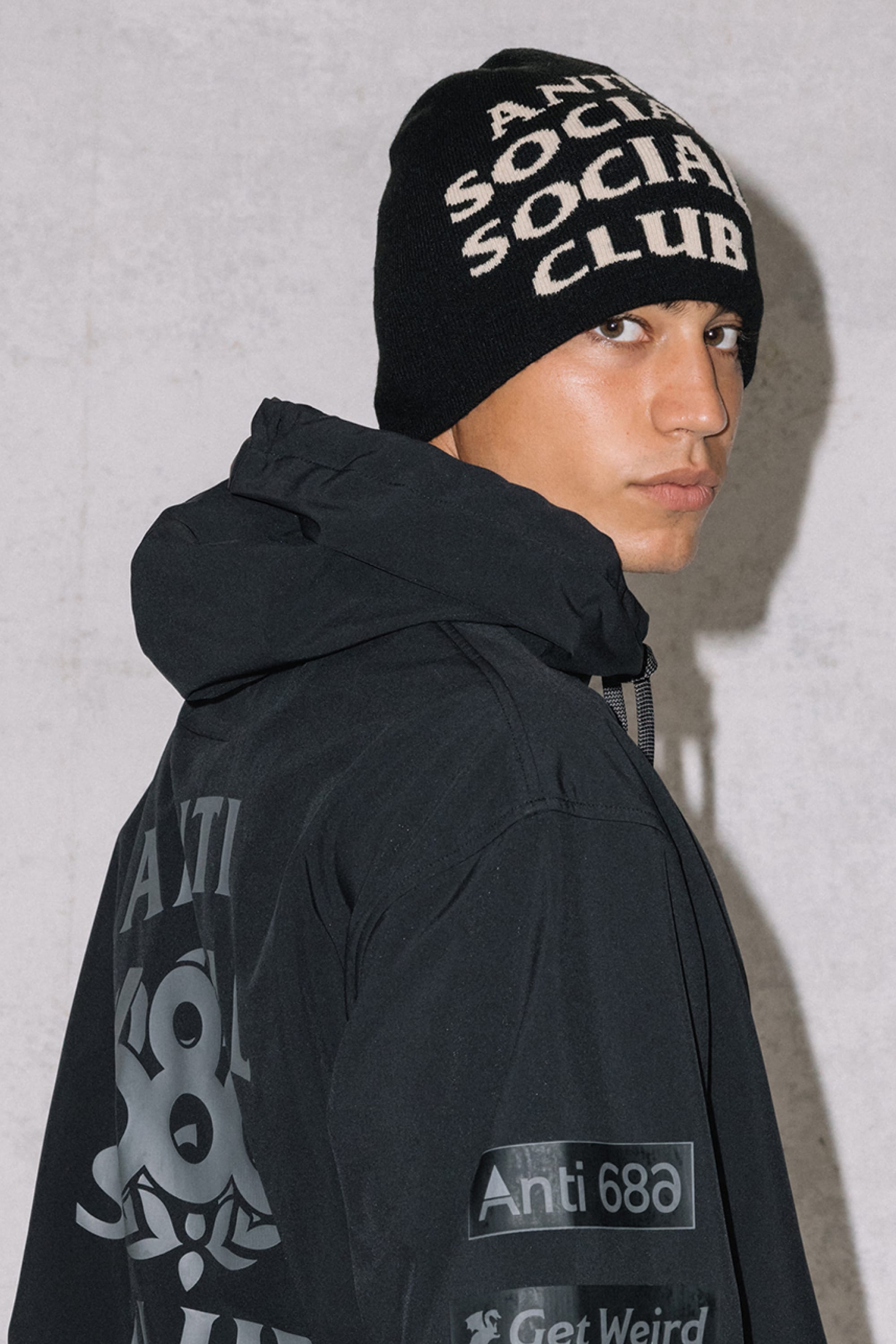 A person wearing a 686 ASSC Skully Beanie by 686, featuring bold ANTI SOCIAL SOCIAL CLUB text, and a black jacket with white lettering, stands against a plain light-colored wall looking over their shoulder at the camera.