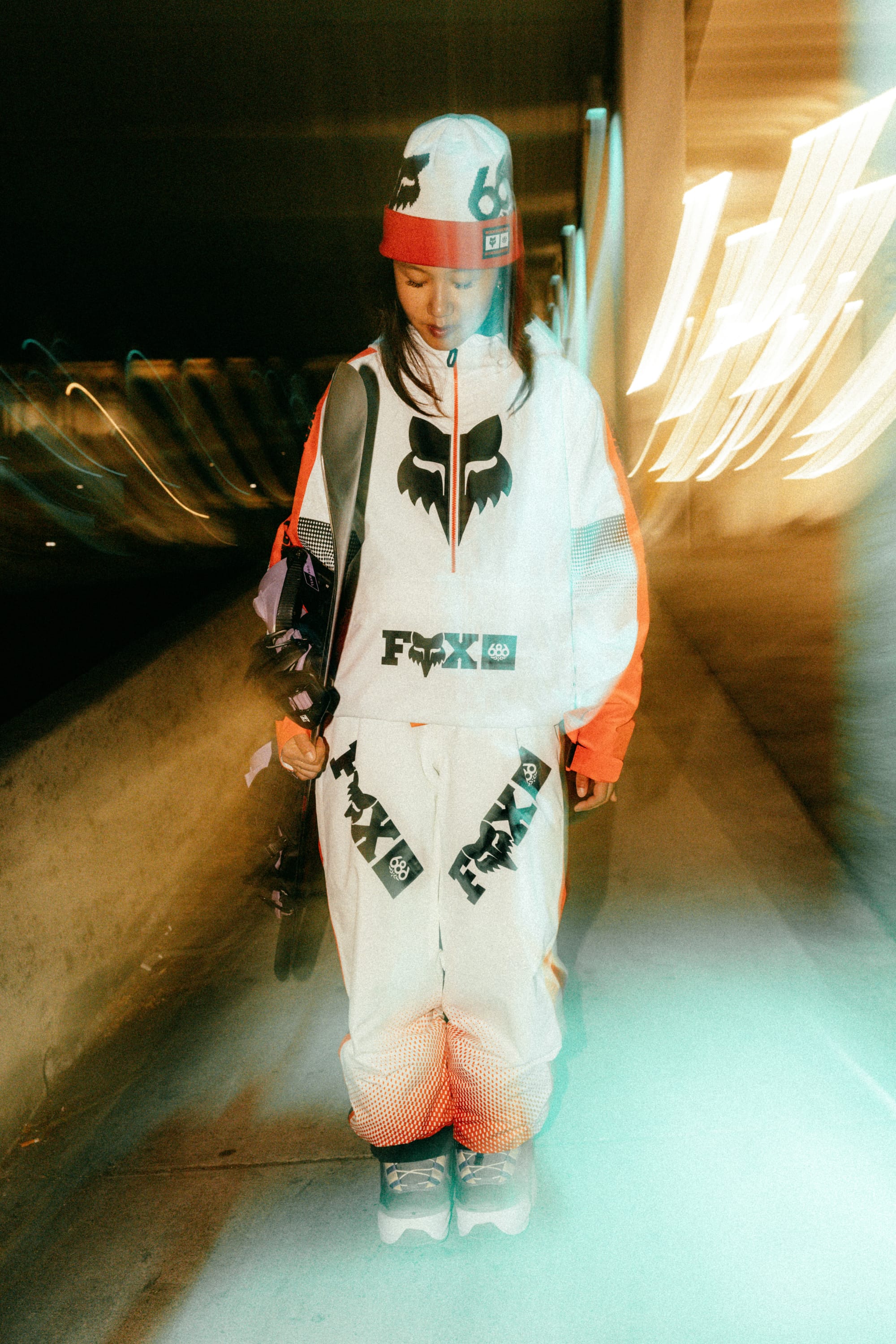 Wearing the 686 Fox Racing Cuff Beanie, a person in a white and orange racing suit with bold black fox logos stands on a city sidewalk at night, holding a skateboard against a backdrop of blurred yellow lights.