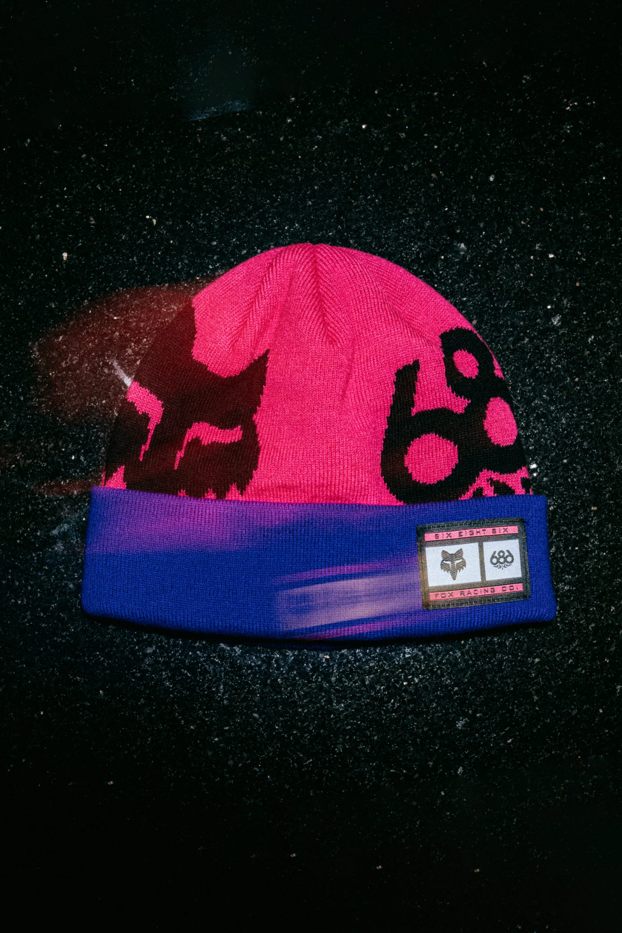 The 686 Fox Racing Cuff Beanie by 686 is a pink beanie with black abstract face designs, a blue brim, and a rectangular patch with symbols and text, shown on a textured black surface.