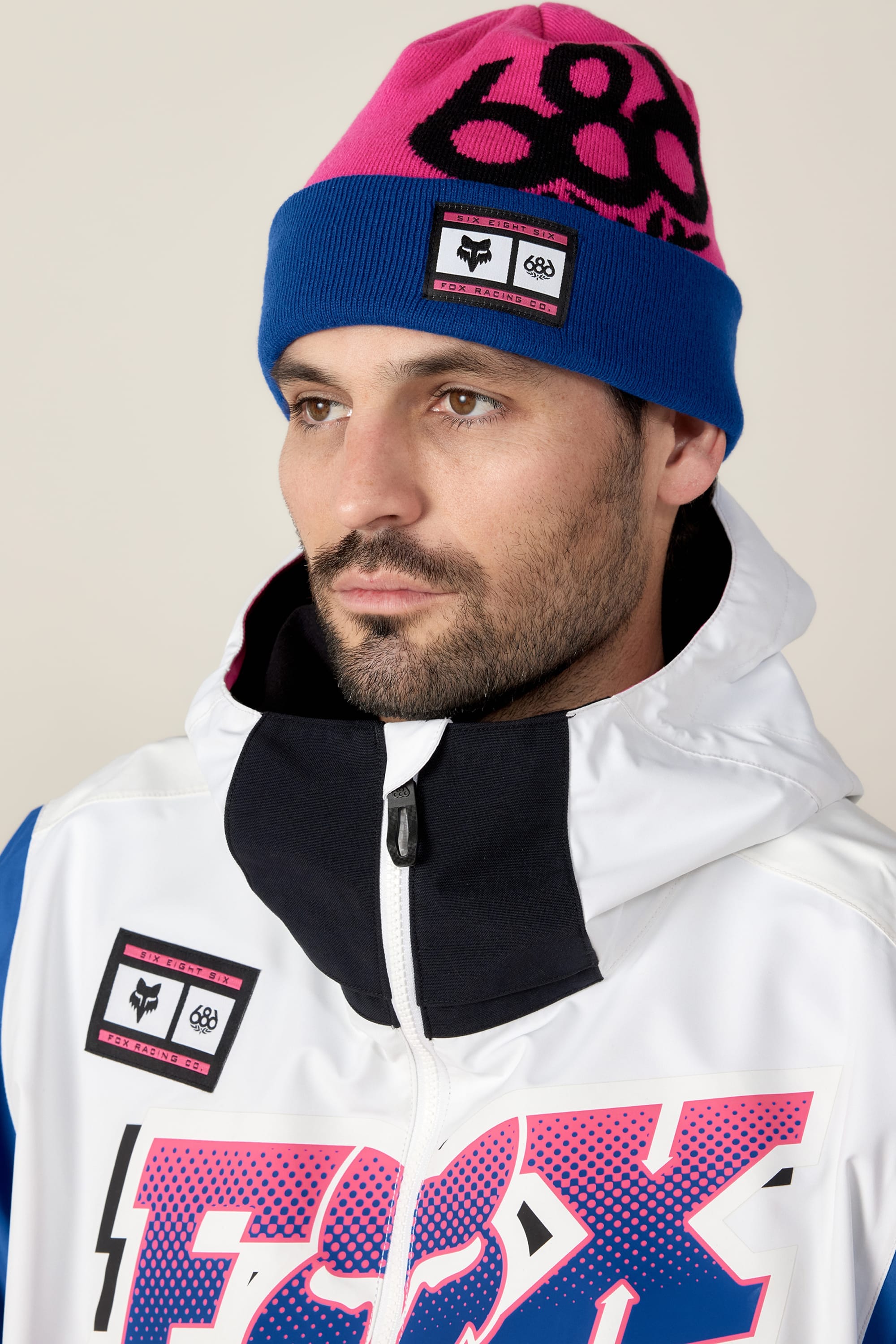 A bearded man wears the 686 Fox Racing Cuff Beanie in pink and blue with a black "88" design, paired with a white jacket featuring a pink and blue FOX logo and a black collar. He looks slightly to the side against a plain background.