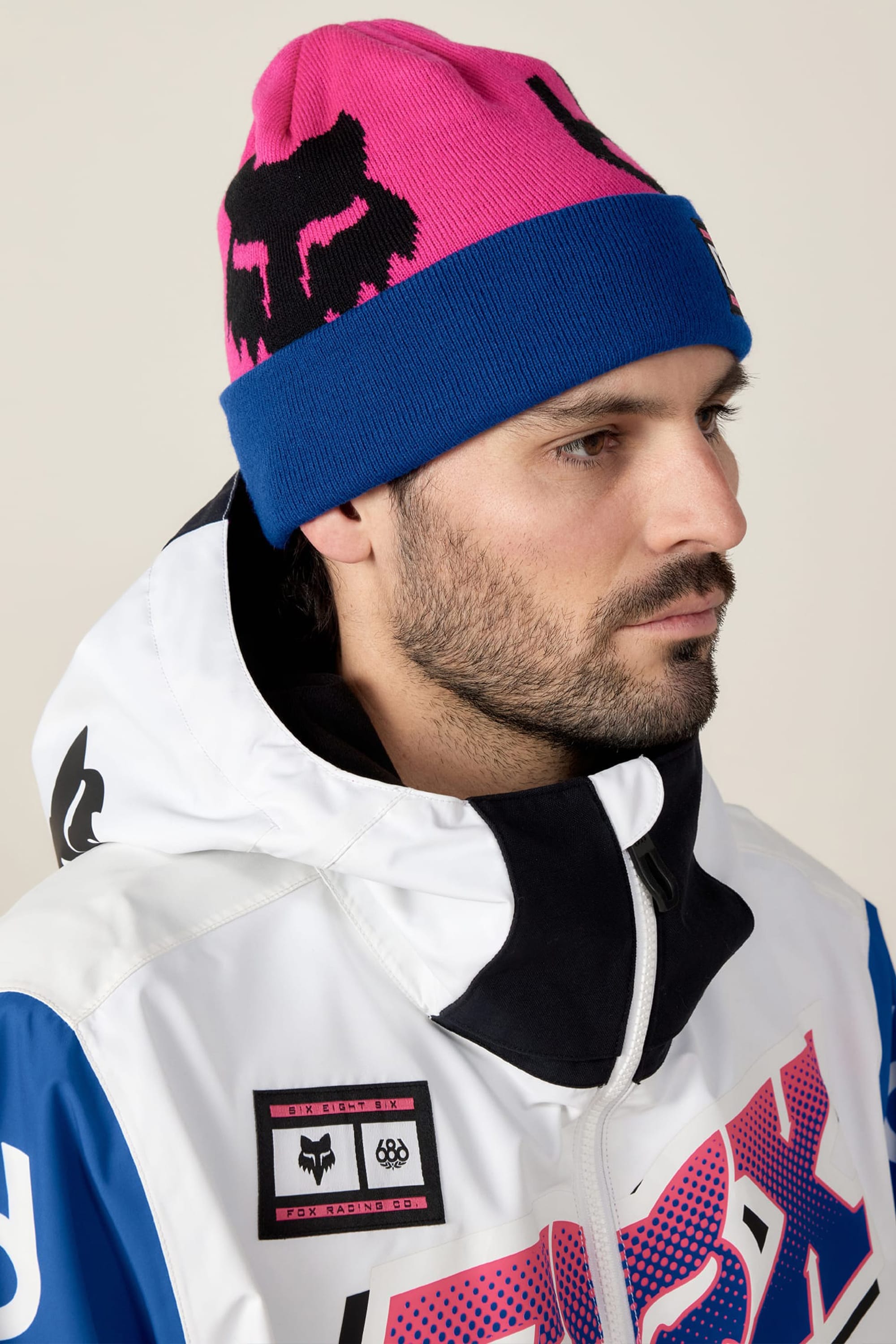 A man with a short beard wears the 686 Fox Racing Cuff Beanie by 686 in pink and blue with a black logo, and a white, blue, and black jacket featuring patches and bold lettering. He looks to the side against a plain background.