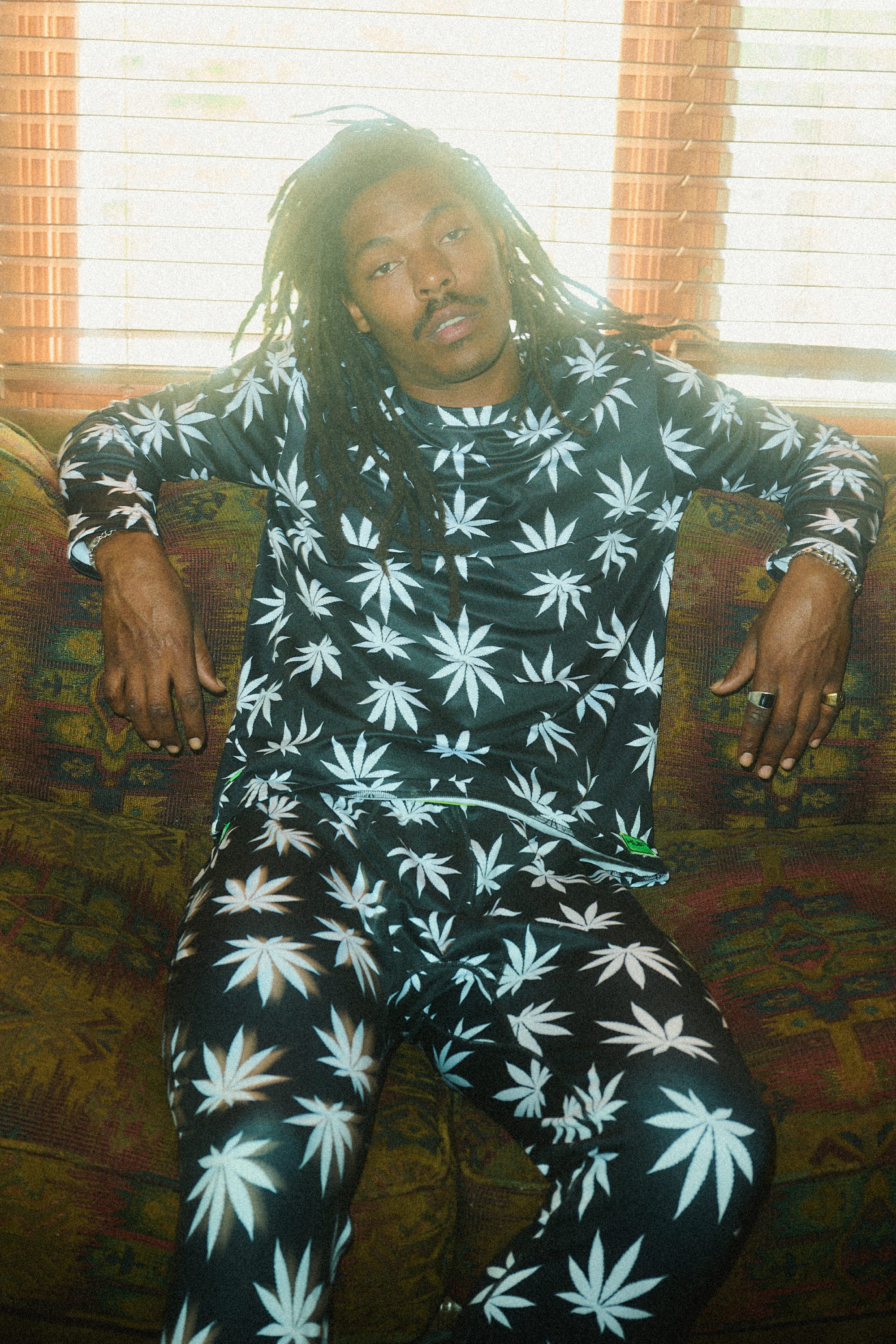 A person with long dreadlocks sits on a patterned couch, wearing the 686 Men's HUF Plantlife Base Layer LS Top by 686, which features large white marijuana leaf prints. Sunlight filters through wooden blinds behind them.