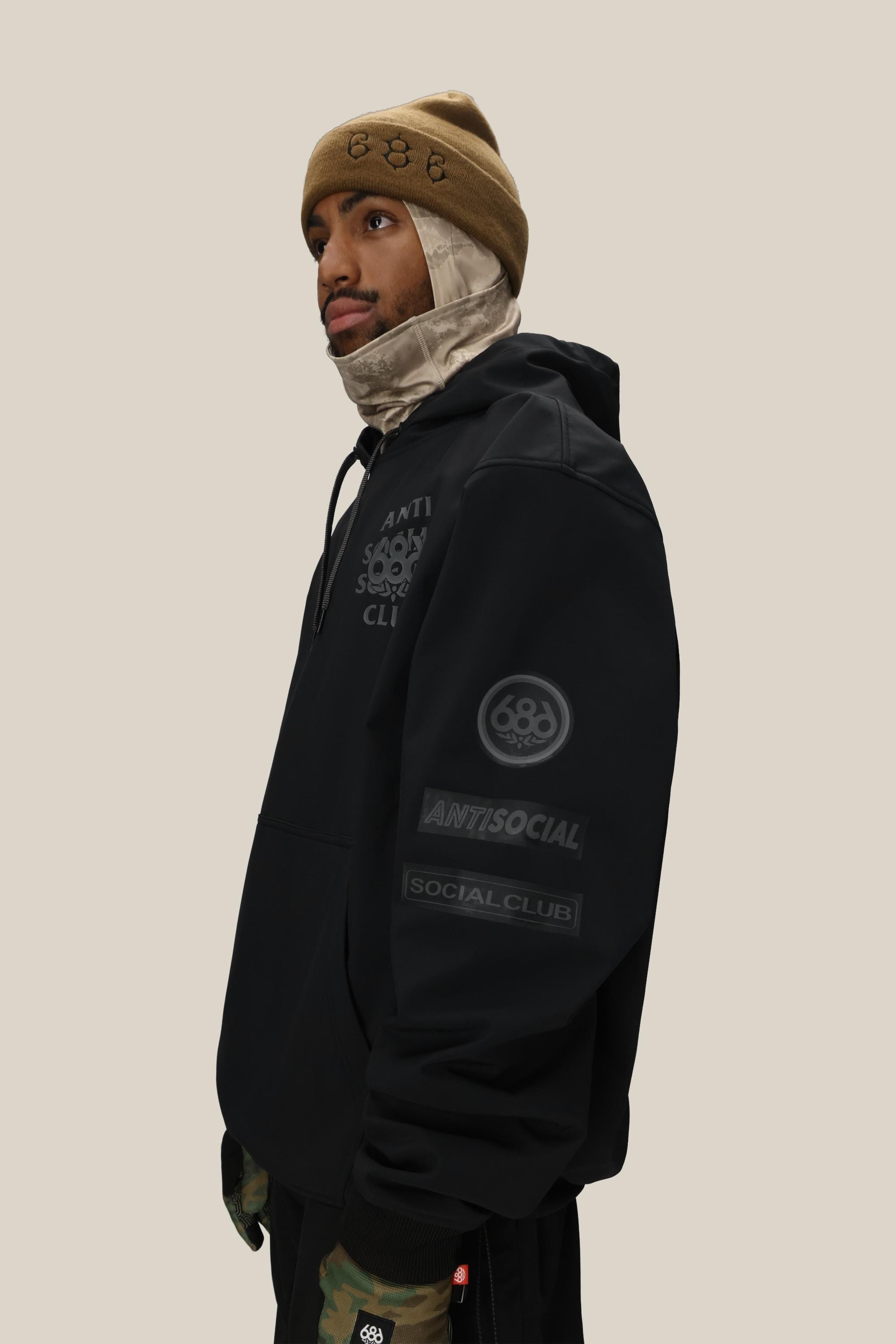 A person in a tan beanie, beige balaclava, and black 686 Men's ASSC F1 Waterproof Team Hoody stands against a plain beige background, looking to the side.