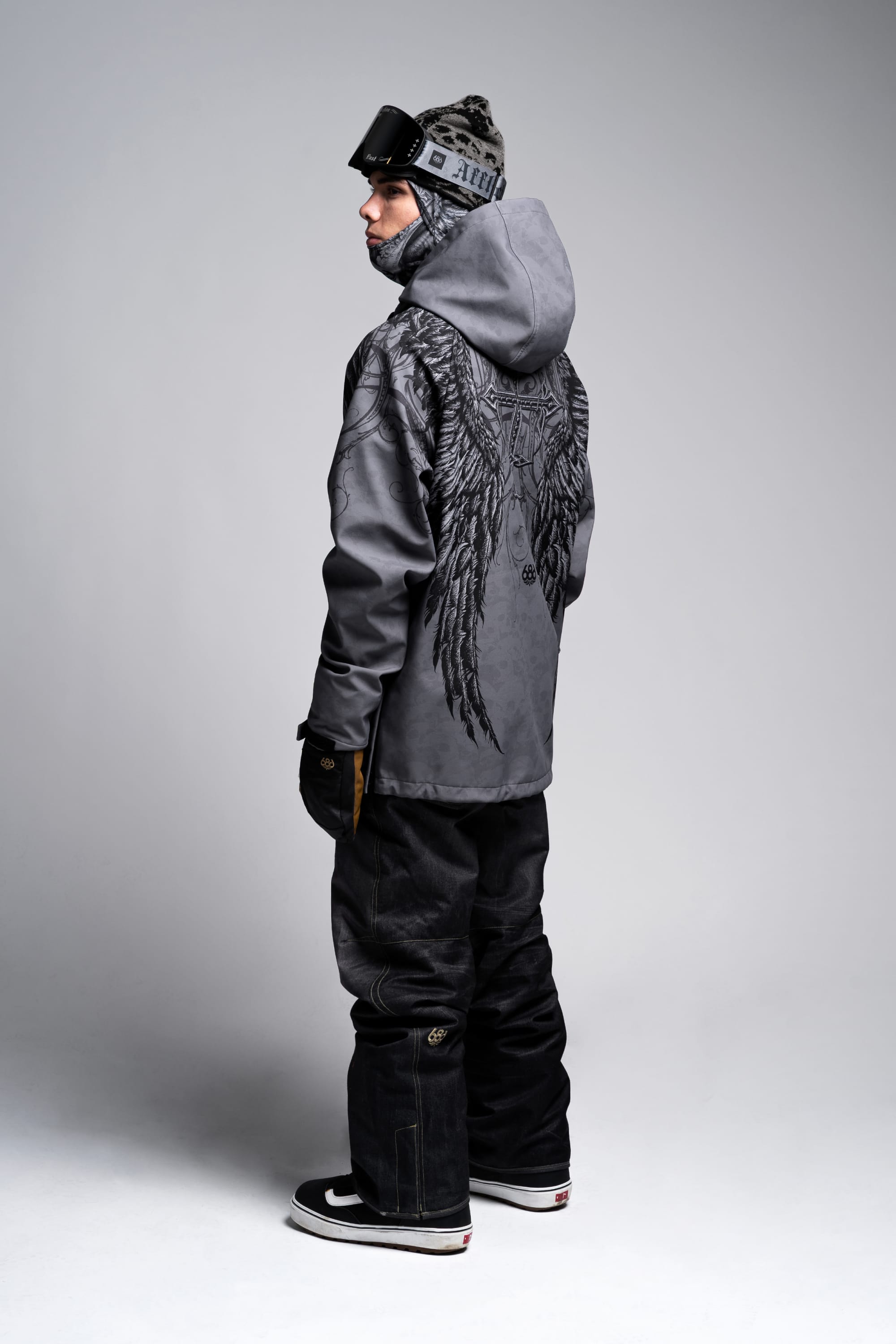 A person in winter gear stands sideways, wearing the 686 Affliction Waterproof Hoody by 686 with a bold black wing design on the back, paired with black pants, gloves, a gray beanie, and ski goggles.