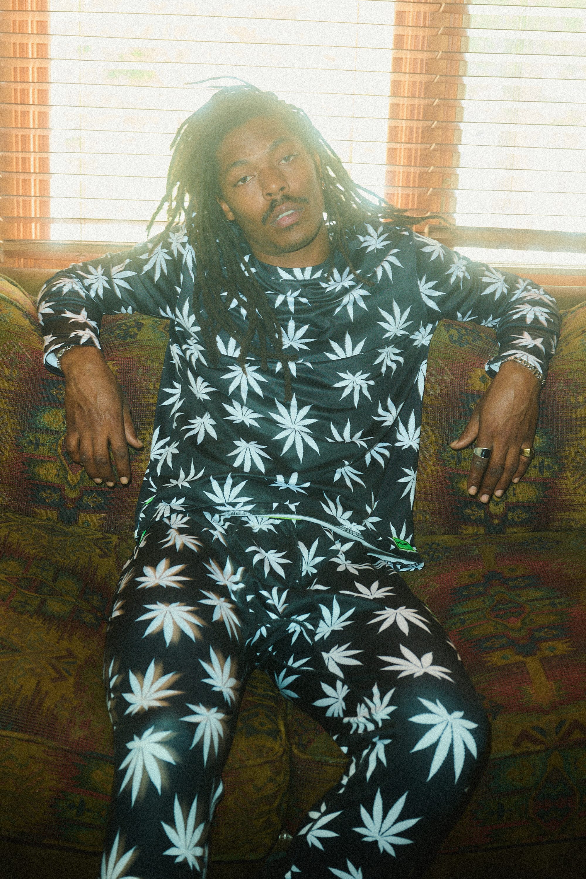 A man with long dreadlocks relaxes on a patterned couch wearing 686 Men's HUF Plantlife Base Layer Bottoms by 686, featuring a black design with white marijuana leaf prints. Light streams in through blinds behind him as he rests his arms on the couch.