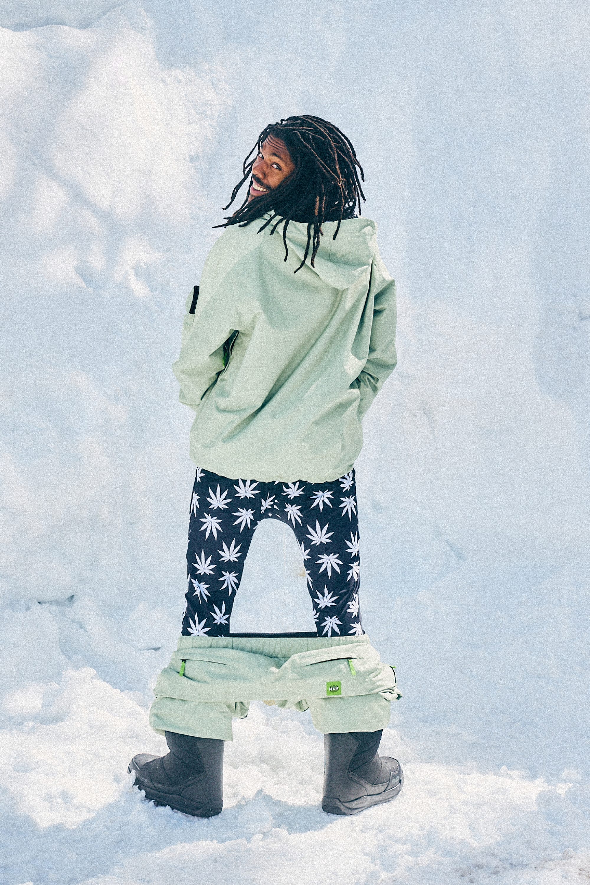 A person stands in the snow with their 686 Men's HUF Plantlife Base Layer Bottom—black leggings with white marijuana leaf patterns—visible under their snow pants. They wear a light green jacket, black boots, and smile over their shoulder.
