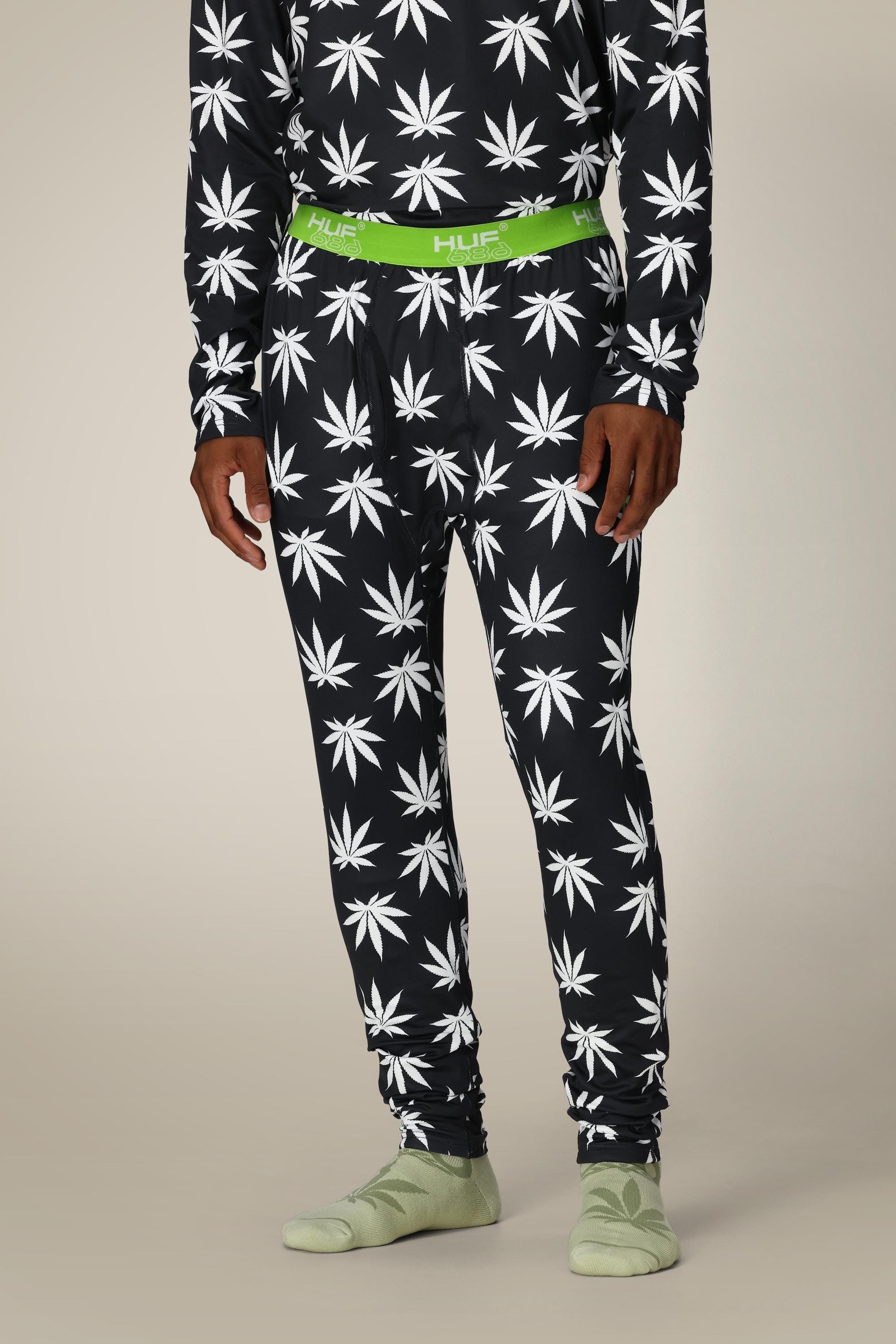 A person models the 686 Men’s HUF Plantlife Base Layer Bottom—black pajama pants with a white cannabis leaf print, green HUF waistband, and light green socks—against a plain beige background. The person’s head is not shown.