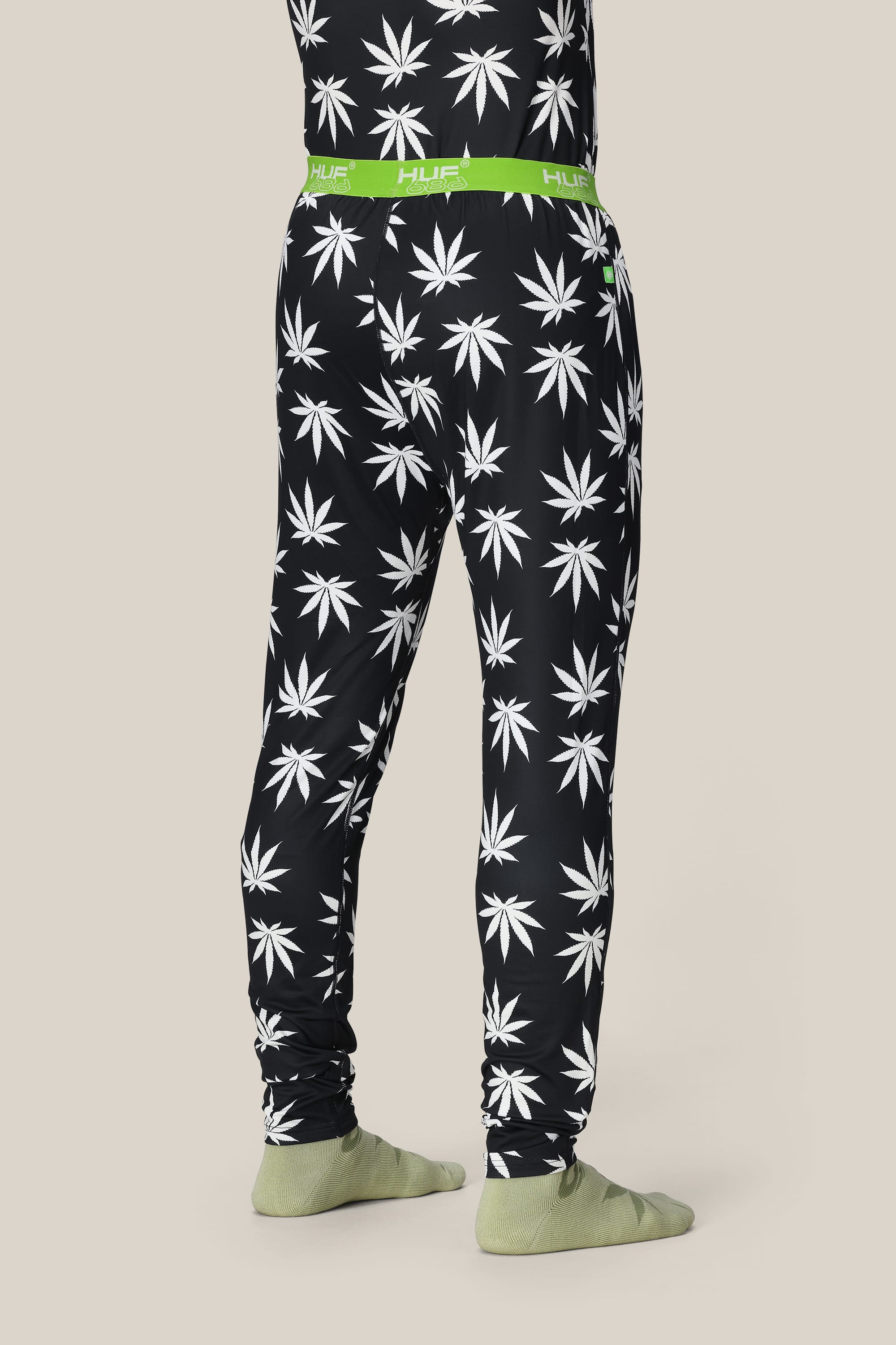 A person wears the 686 Men's HUF Plantlife Base Layer Bottom featuring a black base, white cannabis leaf pattern, and bright green HUF waistband from brand 686, paired with light green socks against a beige background.